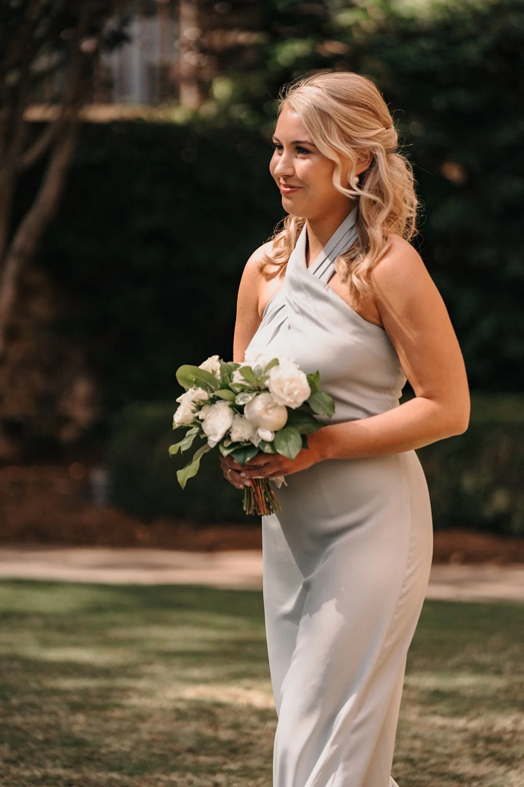 A woman with blonde hair styled in loose curls smiling while holding a bouquet of white flowers and greenery, wearing a light grey halter dress outdoors.