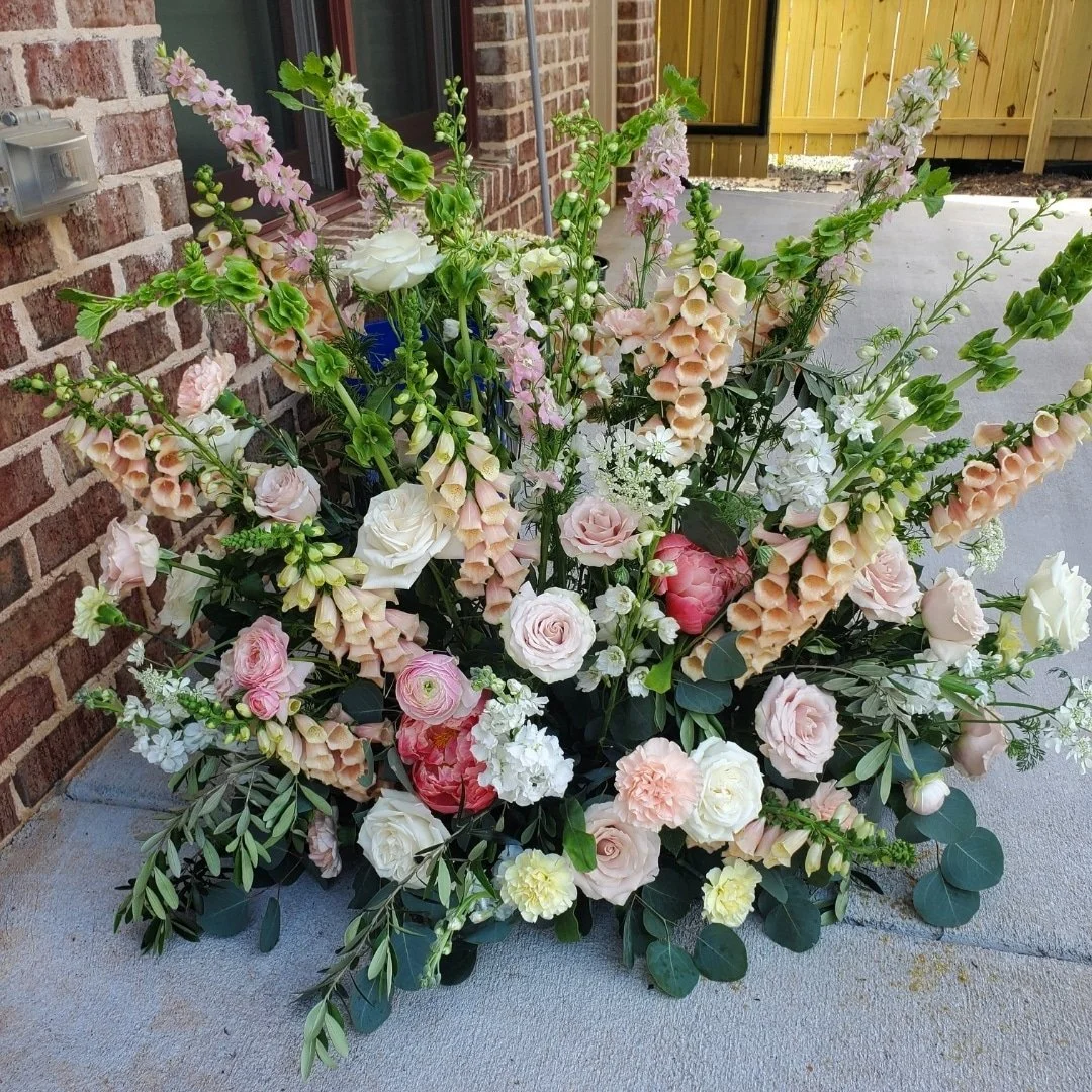 A large, colorful floral arrangement featuring roses, peonies, and snapdragons on a concrete patio next to a brick wall and a wooden fence.
