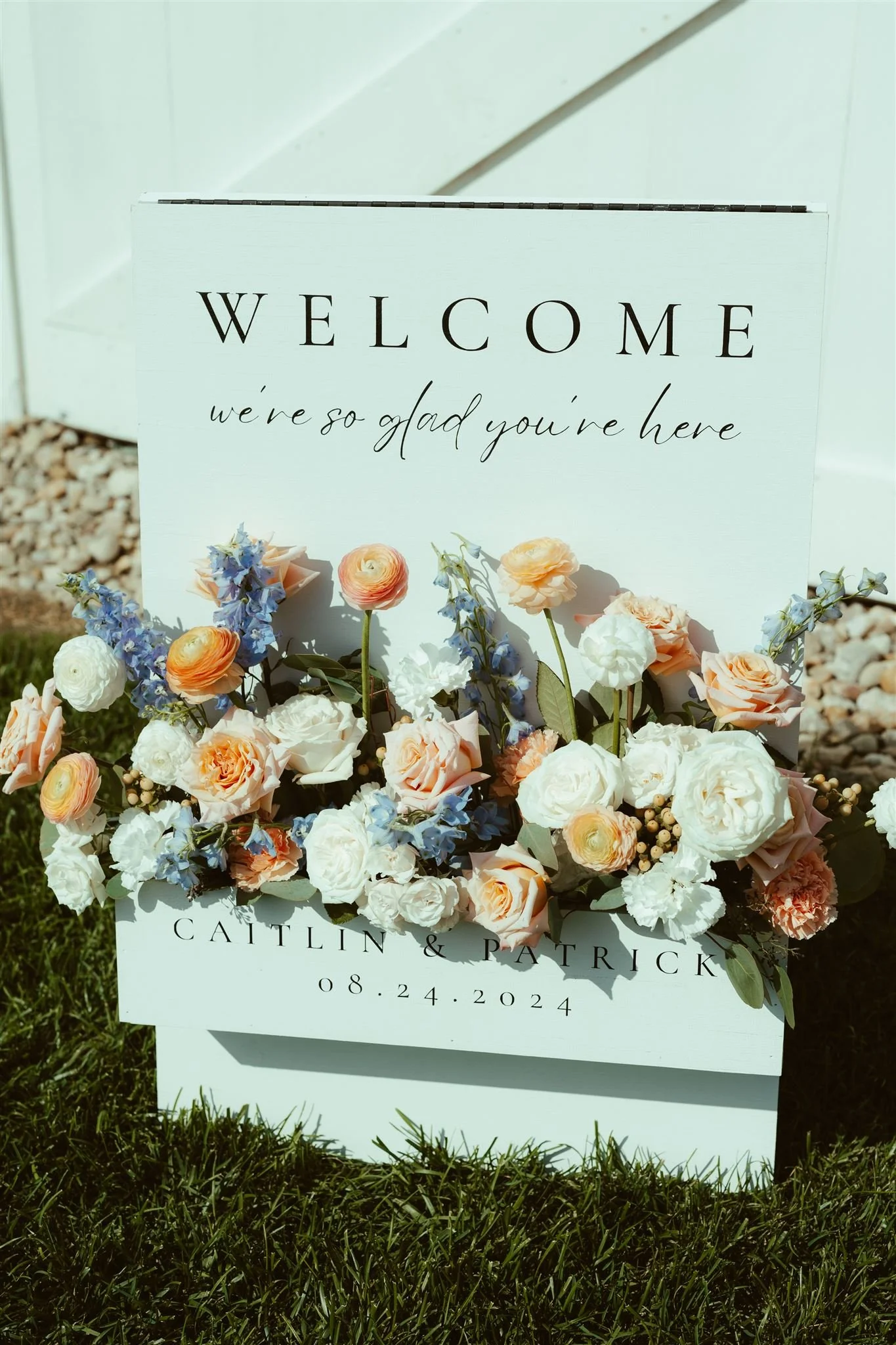 A decorative wooden sign with the text 'WELCOME we're so glad you're here' and 'Caitlin & Patrick 08.24.2024' surrounded by peach, white, and purple flowers at a wedding entrance.