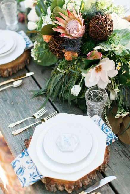 Table setting with white plates, a blue patterned napkin, clear glass, and silver utensils, with a large floral centerpiece featuring pink protea, white magnolia, orange flowers, greenery, and decorative brown woven balls on a rustic wooden table.