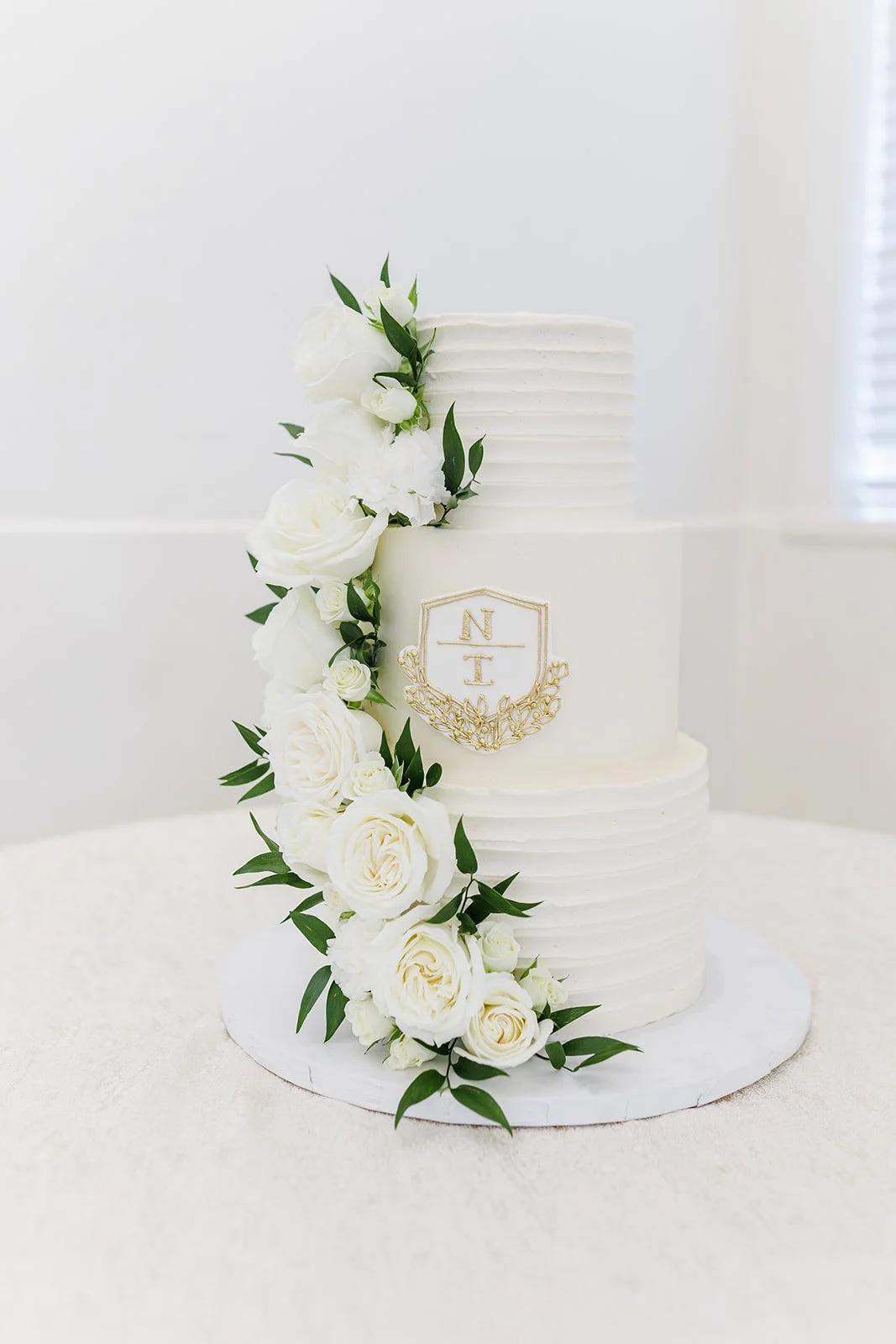 White wedding cake decorated with white roses and greenery, featuring a gold monogram emblem with the initials N and T.