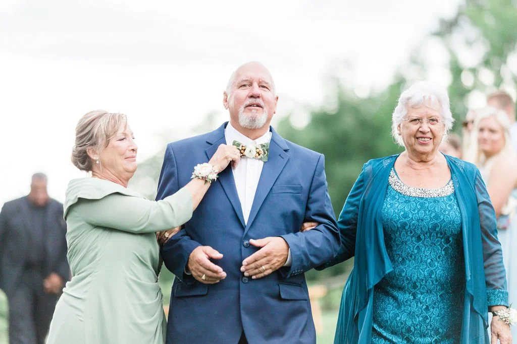 Man in a blue suit being assisted by two women, one on each side, at an outdoor event.