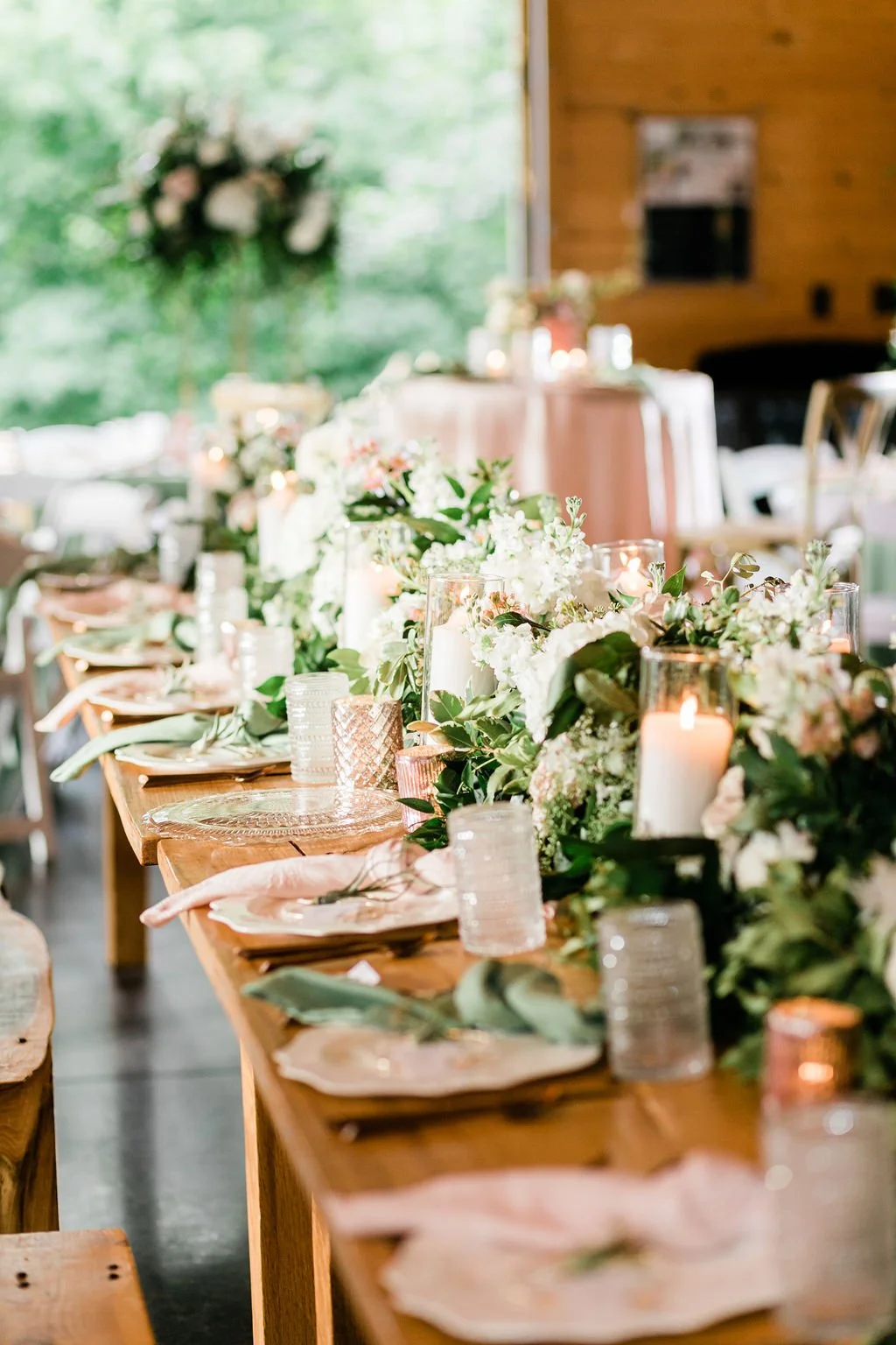 A long wooden dining table decorated with floral arrangements, candles, and elegant tableware. The background shows a window with greenery outside and a wooden interior wall.