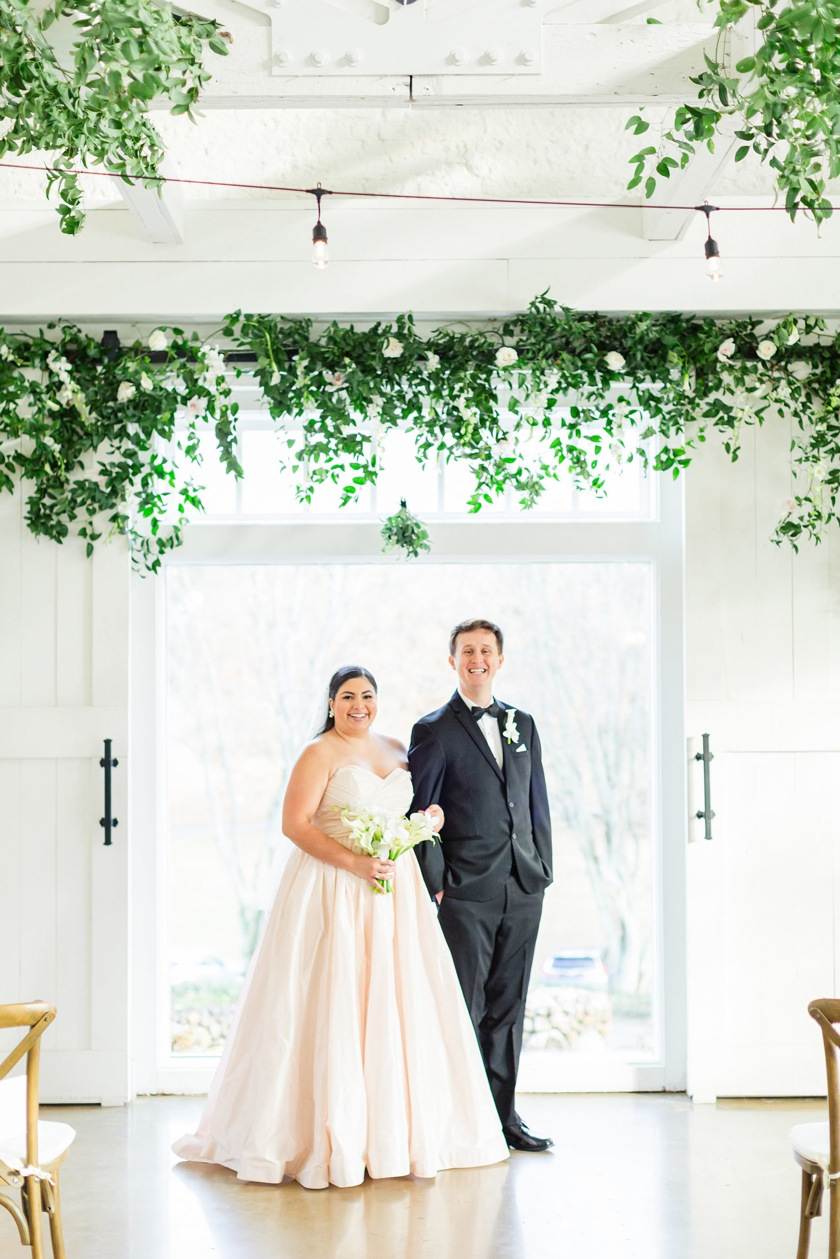 Bride and groom standing inside a decorated venue with greenery and string lights, smiling.