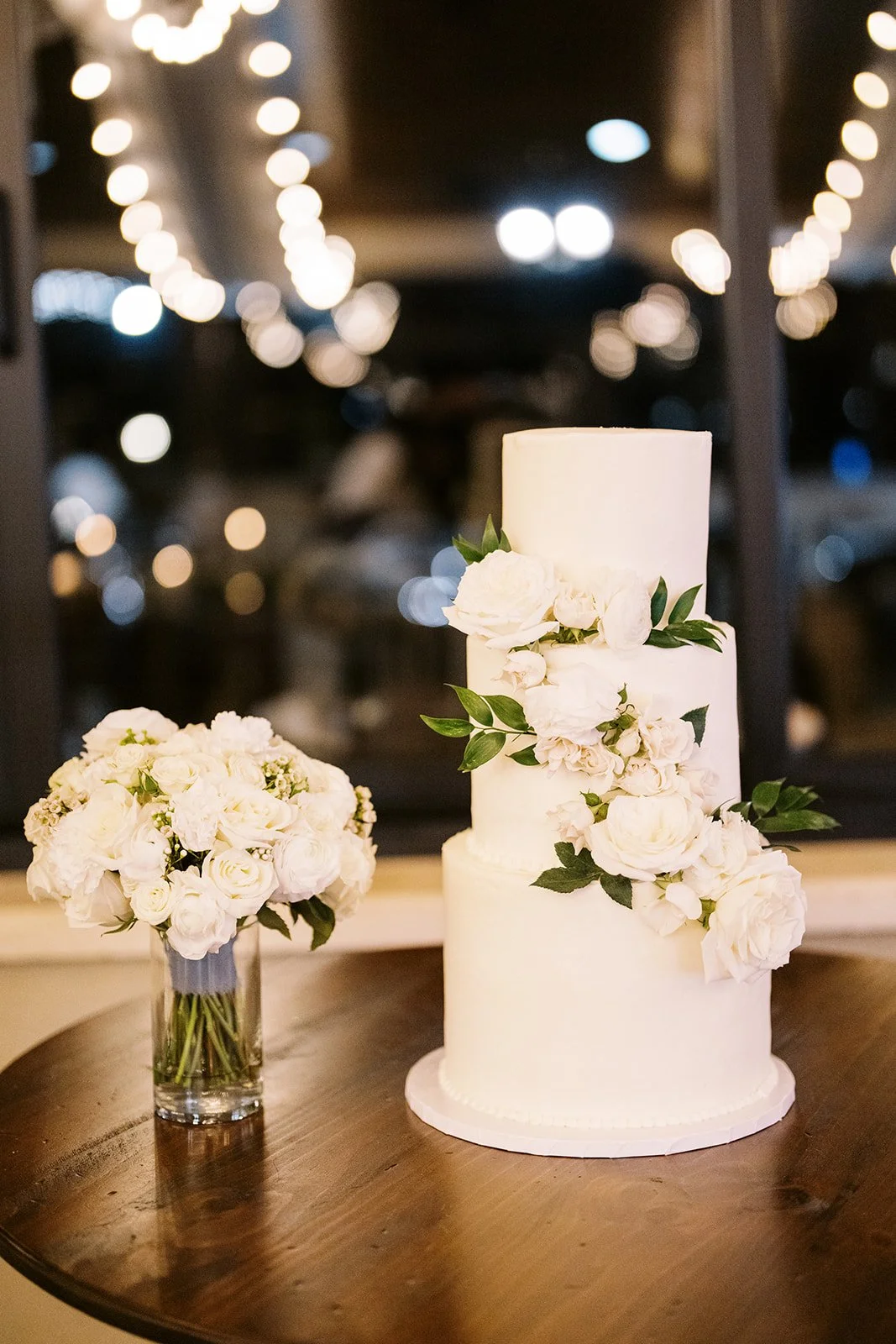 White three-tier wedding cake decorated with white flowers and green leaves on a wooden table, with a bouquet of white flowers in a glass vase beside it.
