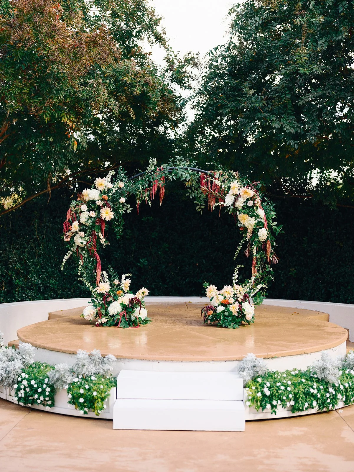 Decorated circular platform with floral arrangements in front of a backdrop of trees, likely for a wedding or outdoor event.