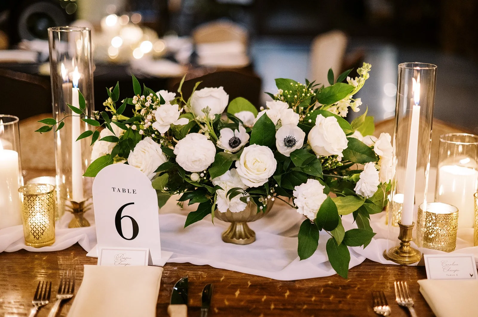 Elegant table centerpiece with white flowers and greenery, surrounded by tall glass candlesticks with white candles and gold votive candle holders, set on a wooden table for a wedding or formal event.