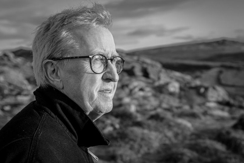 Black and white photo of a man with glasses looking off into the distance with Dartmoor landscape behind.