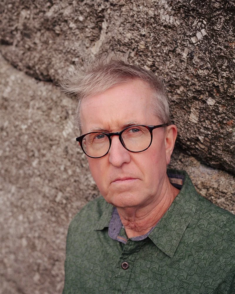 Ian Wellens - A man with glasses, wearing a green patterned shirt, standing outdoors against a rocky background.
