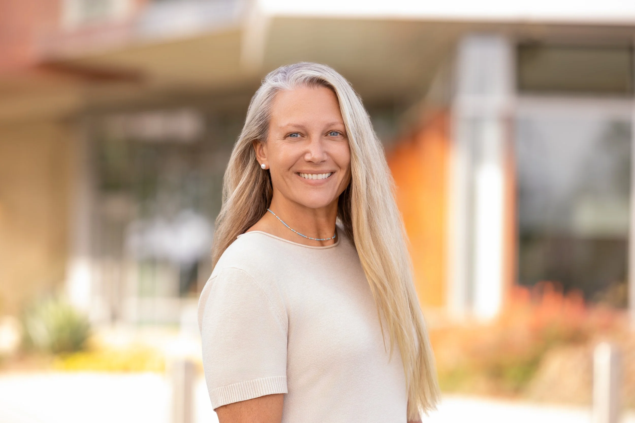 A smiling woman with long blonde hair standing outdoors in front of a blurred modern building background.