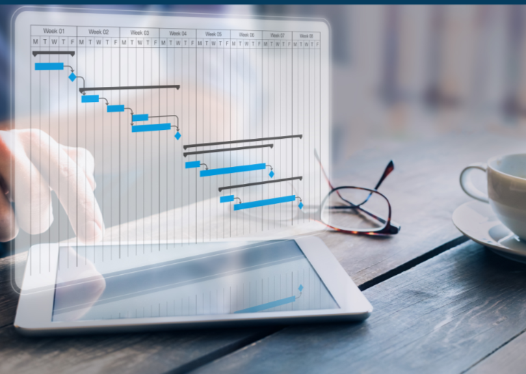 Tablet displaying a digital Gantt chart for project planning, with a person's hand pointing at it, on a wooden table with glasses and a coffee cup nearby.