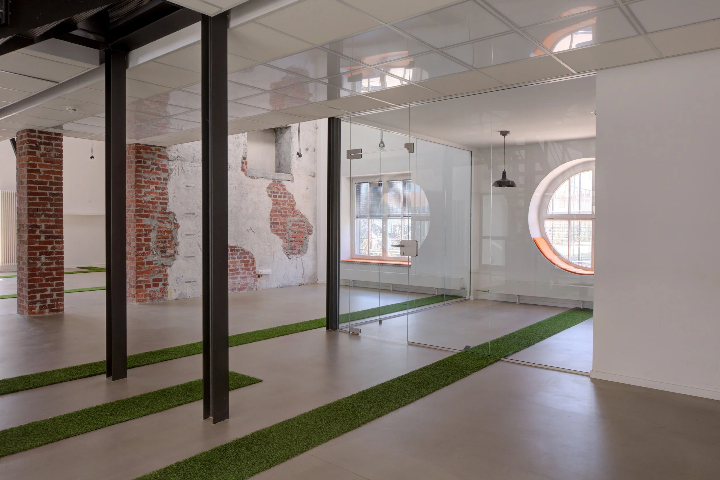 Moderner Büroinnenraum mit Glaswänden, Sicht auf eine Fensterwand mit rundem Fenster und Ziegelwand, helles Design, grüne Teppichstreifen auf glattem Boden.