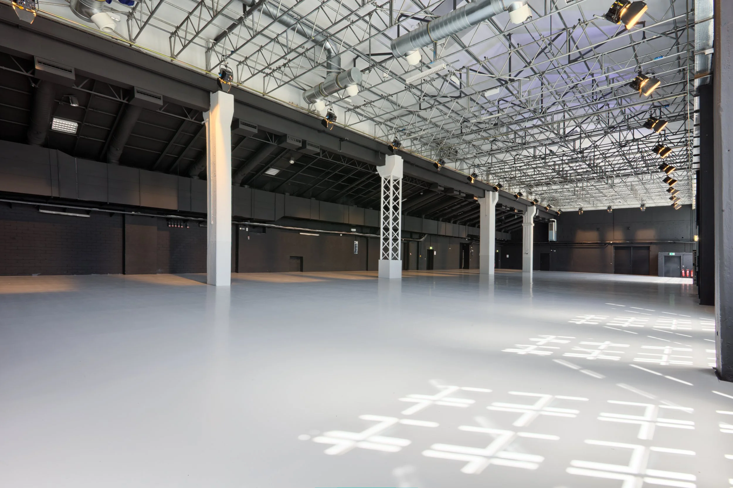 Großer, leerer Indoor-Sporthallenraum mit hellem Boden, schwarzen Wänden und Metallträgern an der Decke, Lichtreflexionen auf dem Boden