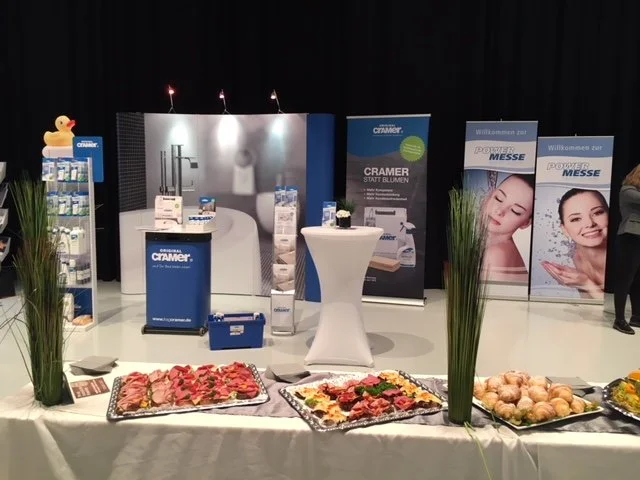 Stand bei einer Messe mit Werbebannern, Verkaufsständen und einem Buffet mit allerlei Häppchen und Punsch. Deko mit Grünpflanzen.