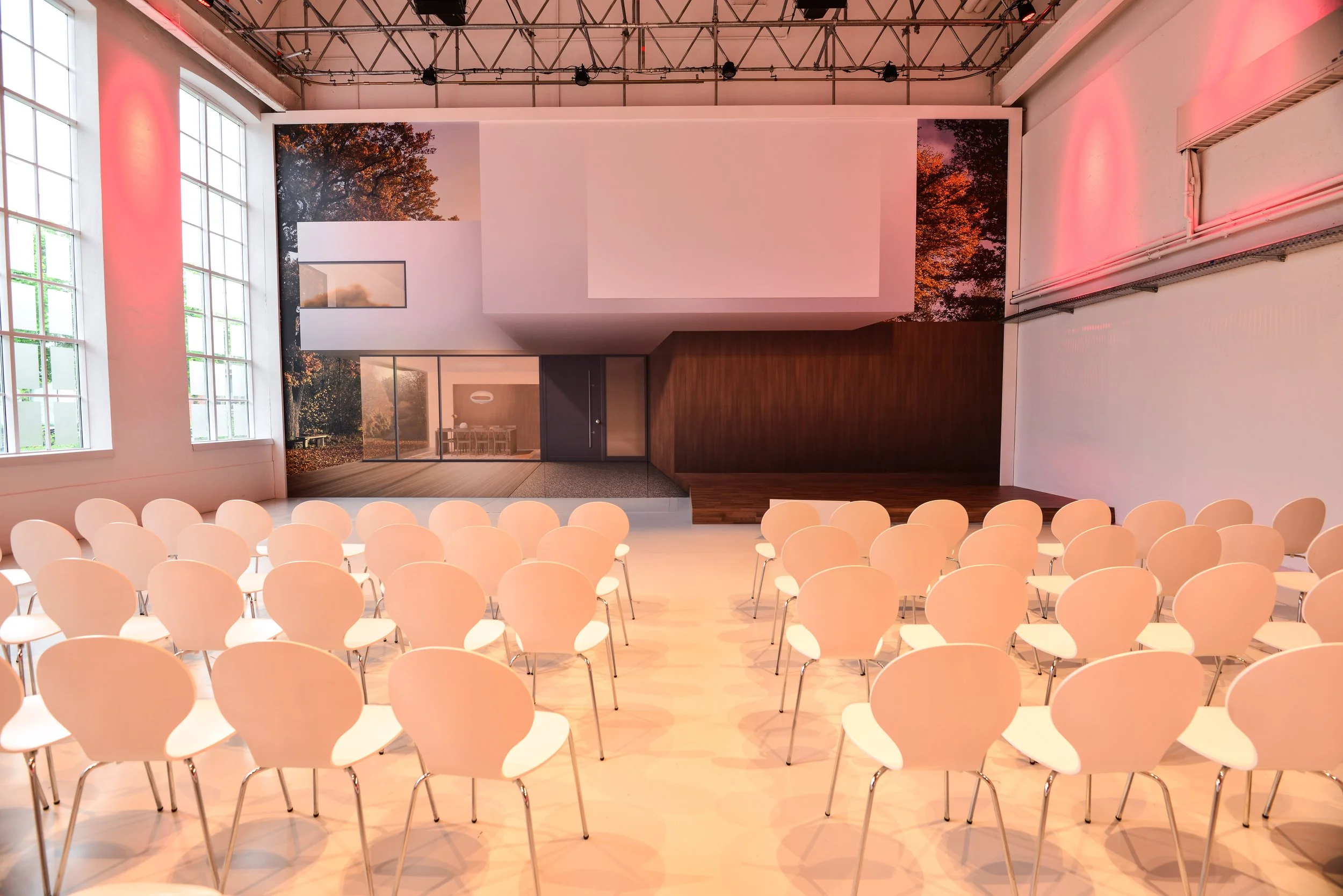 Ein Konferenzraum mit weißen Stühlen, großen Fenstern und einer großen Leinwand an der Wand. Es gibt eine kleine, erhöhte Bühne vorne. Das Licht ist warm und hat einen rosafarbenen Ton.