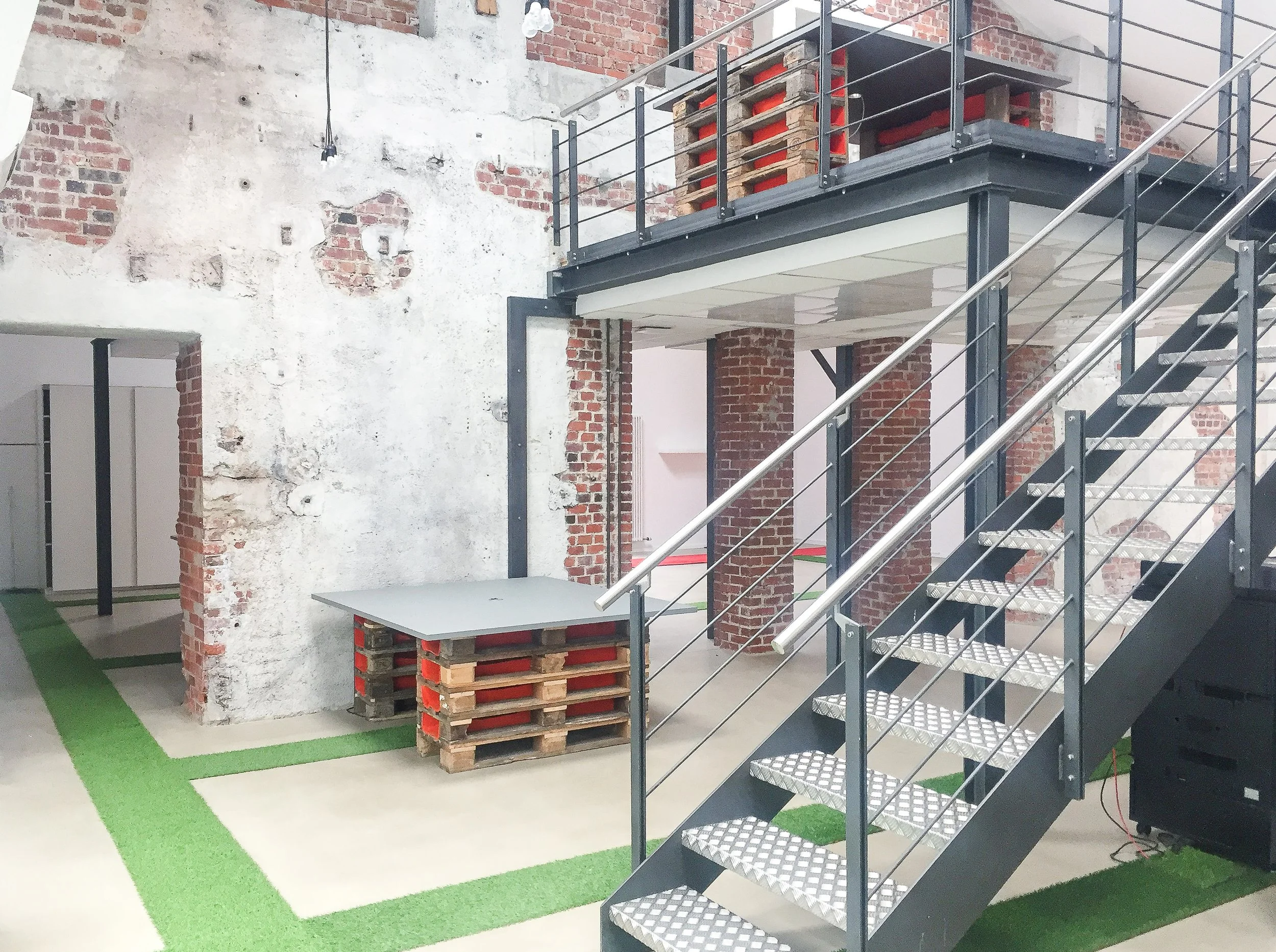 Innenaufnahme eines modernen Bürogebäudes mit offener Architektur, sichtbaren roten Backsteinwänden, einer Metalltreppe mit Geländer, einer Obergeschossplattform und einem Arbeitsplatz aus Palettenholz an der Wand.