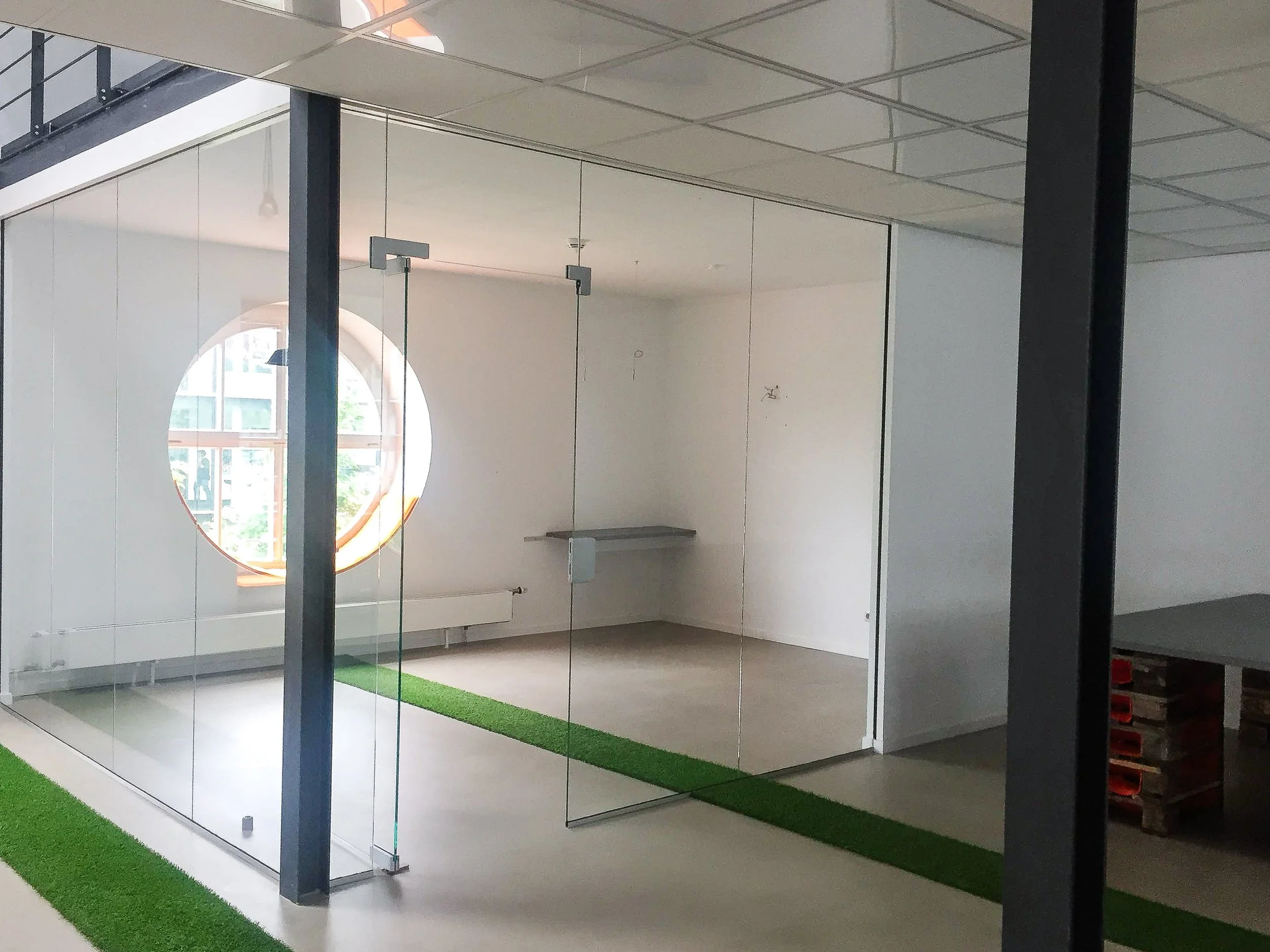 Leerer Raum mit Glaswänden, gelber runder Fenster, graue Teppichböden, grüner Teppichstreifen, und einfache Einrichtung.