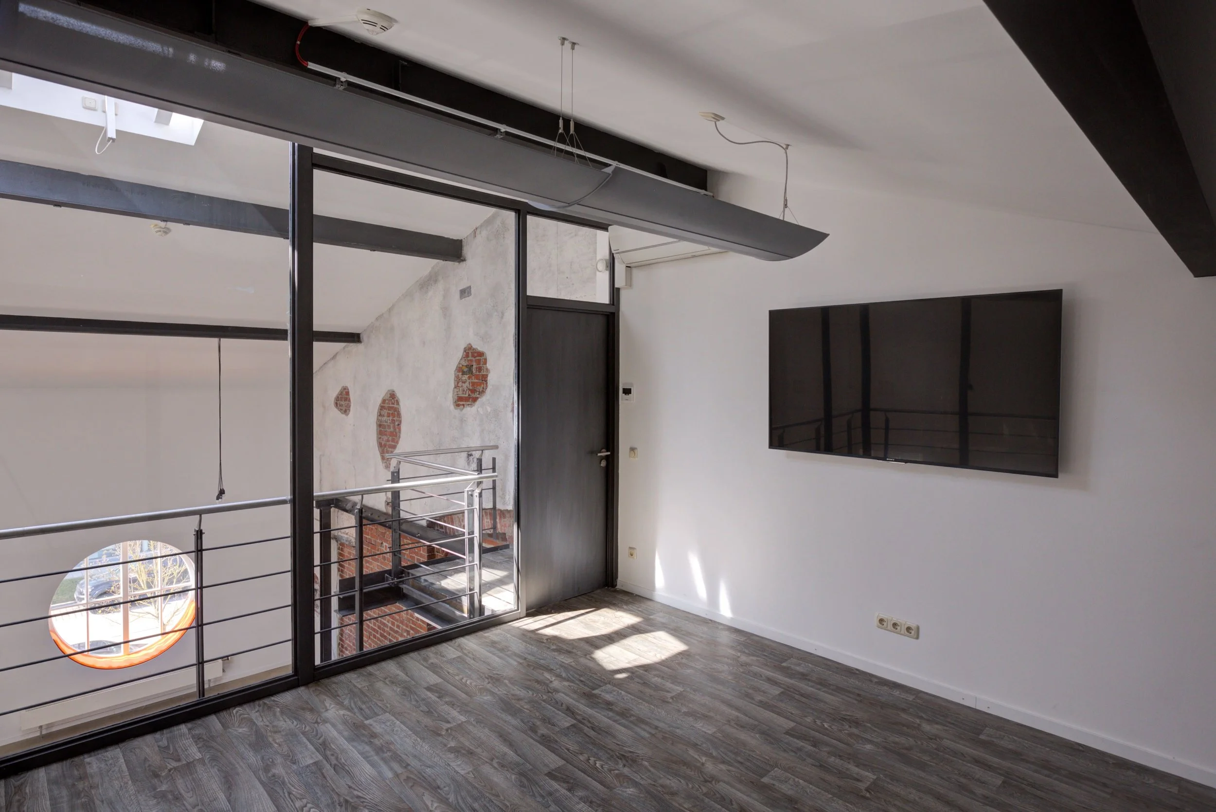 Innenraum eines modernen, unfertigen Apartments mit weißen Wänden, dunklem Laminatboden, einer großen schwarzen Wandversion eines Fernsehers, einer schwarzen Tür, einem offenen Balkon mit Metallgeländer und einer Wand mit abblätternder alter Putzschi