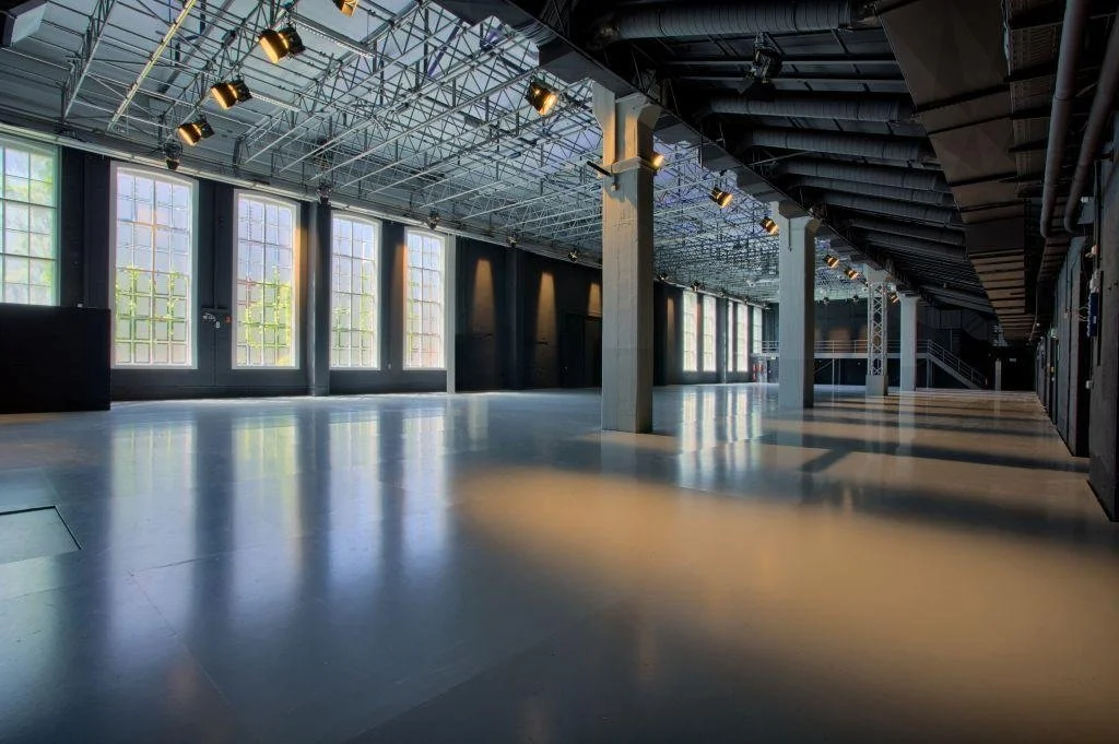 Ein großes, leeres, modern eingerichtetes Event- oder Ausstellungsraum mit hohen Fenstern, Sichtbetonstützen, einer offenen Decke mit sichtbarer Bau- und Lichttechnik und einer glatten, reflektierenden Fliesenboden.