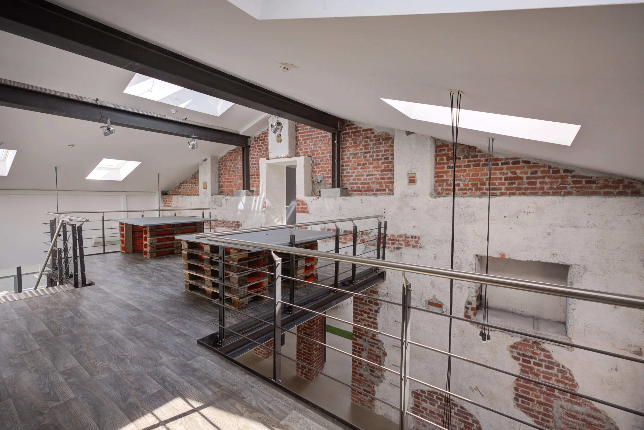 Innenansicht eines modernen Raums im Bau, mit Holzboden, Metallelementen, Fenstern im Dach, unvollendeter Wand mit sichtbarem Ziegel- und Putzmauerwerk, Holzpaletten als Tische, und Bauarbeiten sichtbar.