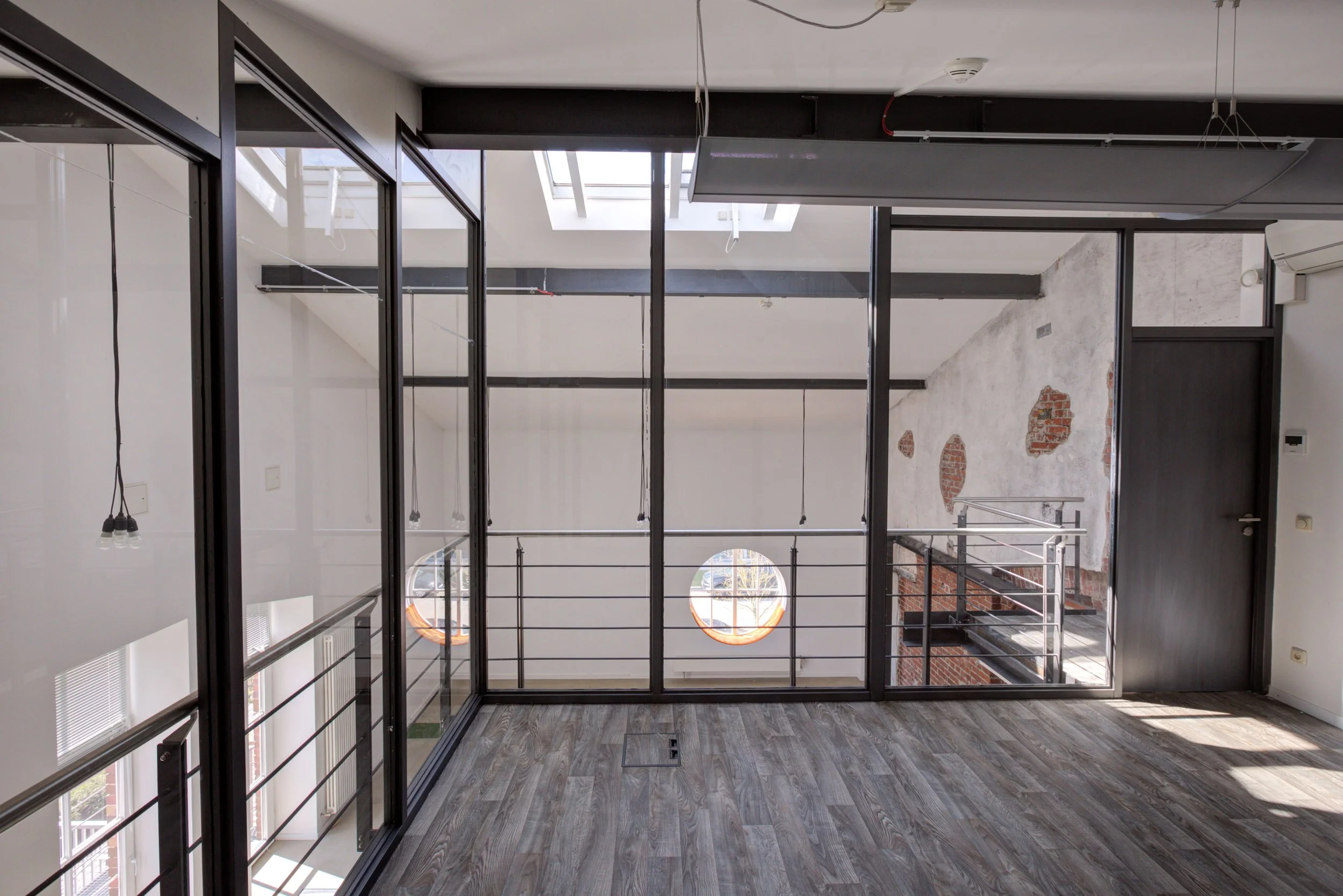 Innenansicht eines modernen, unmöblierten Raums mit großen Glasfenstern, Holzfußboden, einer Deckenfensteröffnung und teilweise sichtbarem rotem Backstein an der Wand.