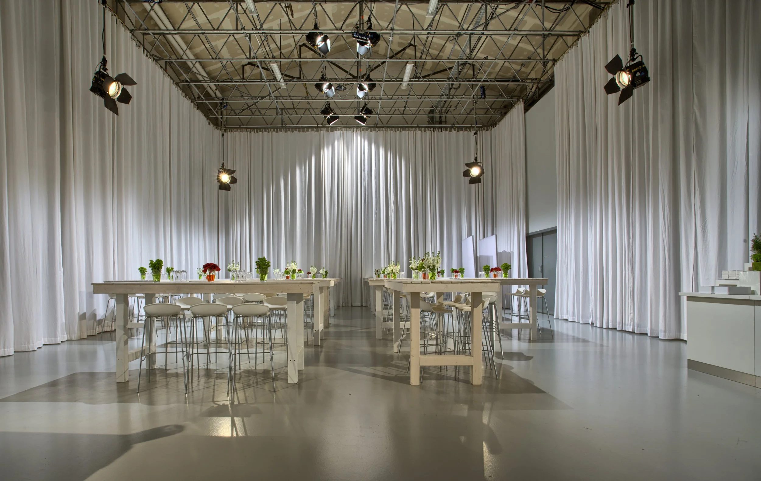 Eleganter Veranstaltungsraum mit weißen Vorhängen, Tischen mit weißen Oberflächen, bunten Blumenarrangements und Stühlen, gut beleuchtet mit Decken- und Wandflutern