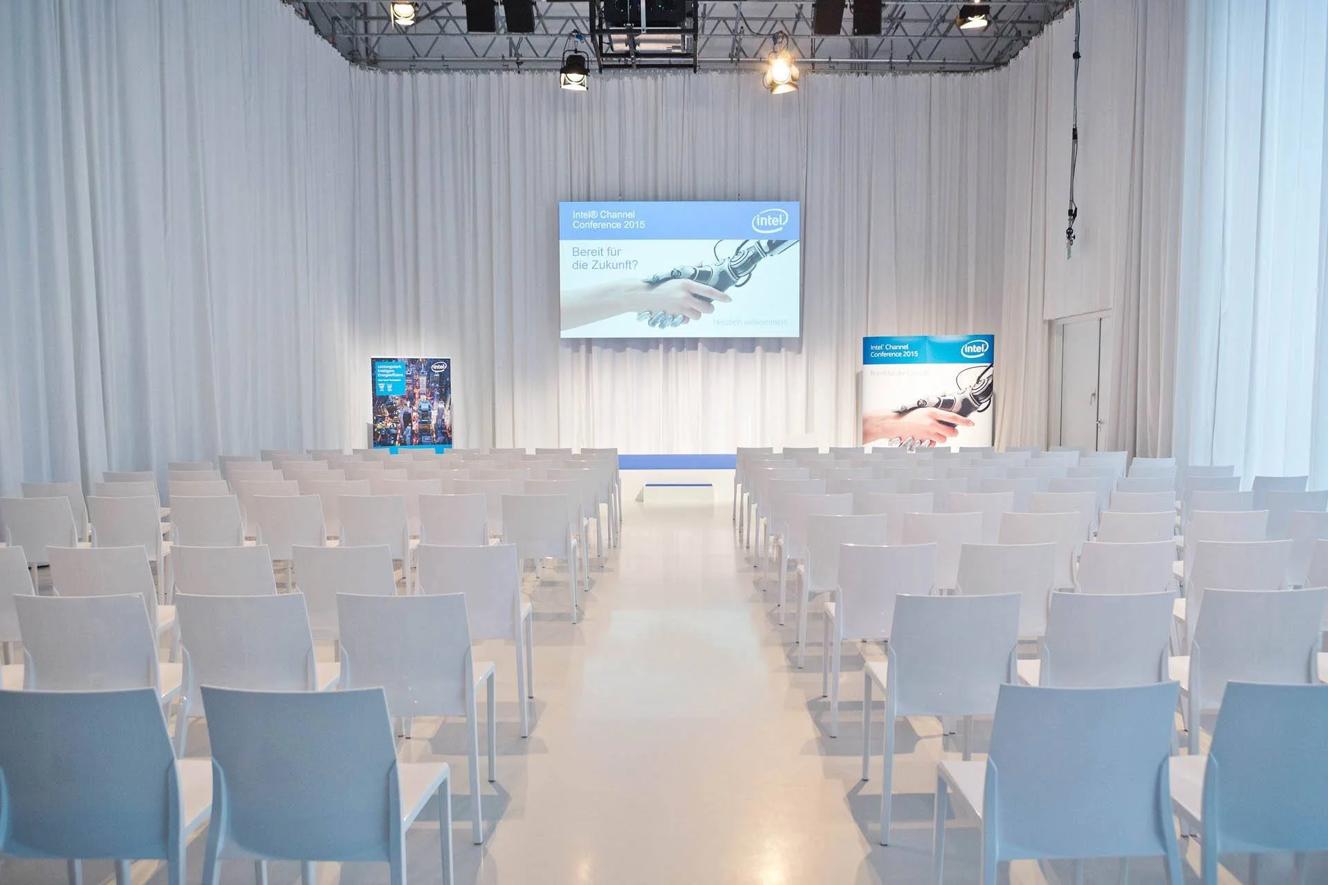 Leerer Konferenzraum mit weißen Stühlen, einer großen Projektionswand mit dem Intel-Logo und Text, und weißen Vorhängen im Hintergrund.