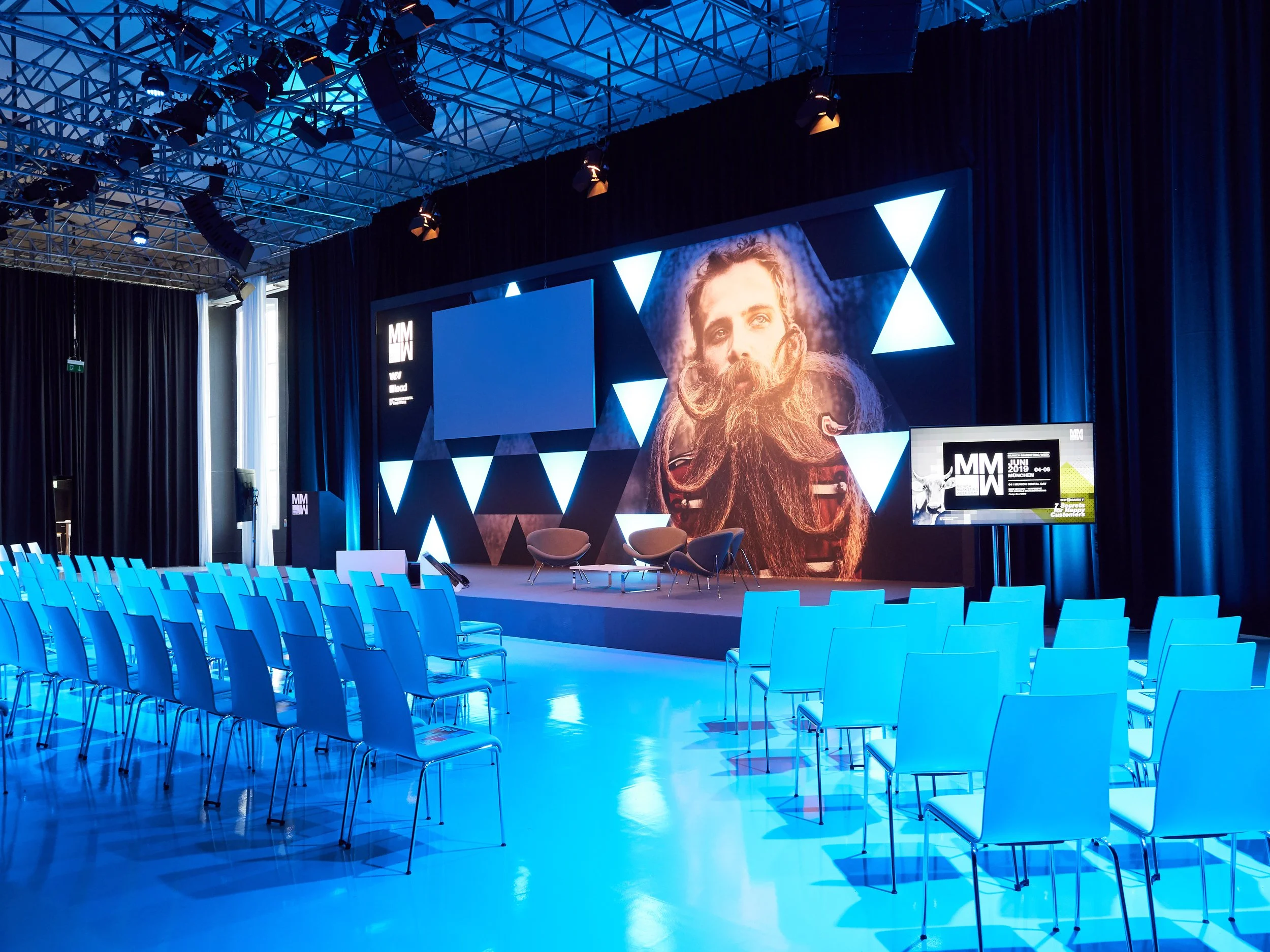 Leerer Konferenzraum mit blauen Stühlen, einer großen Bühne, einem großen Bildschirm mit einem Portrait eines Mannes und mehreren kleineren Bildschirmen, dunklen Vorhängen und einer gewölbten Decke mit Scheinwerfern.