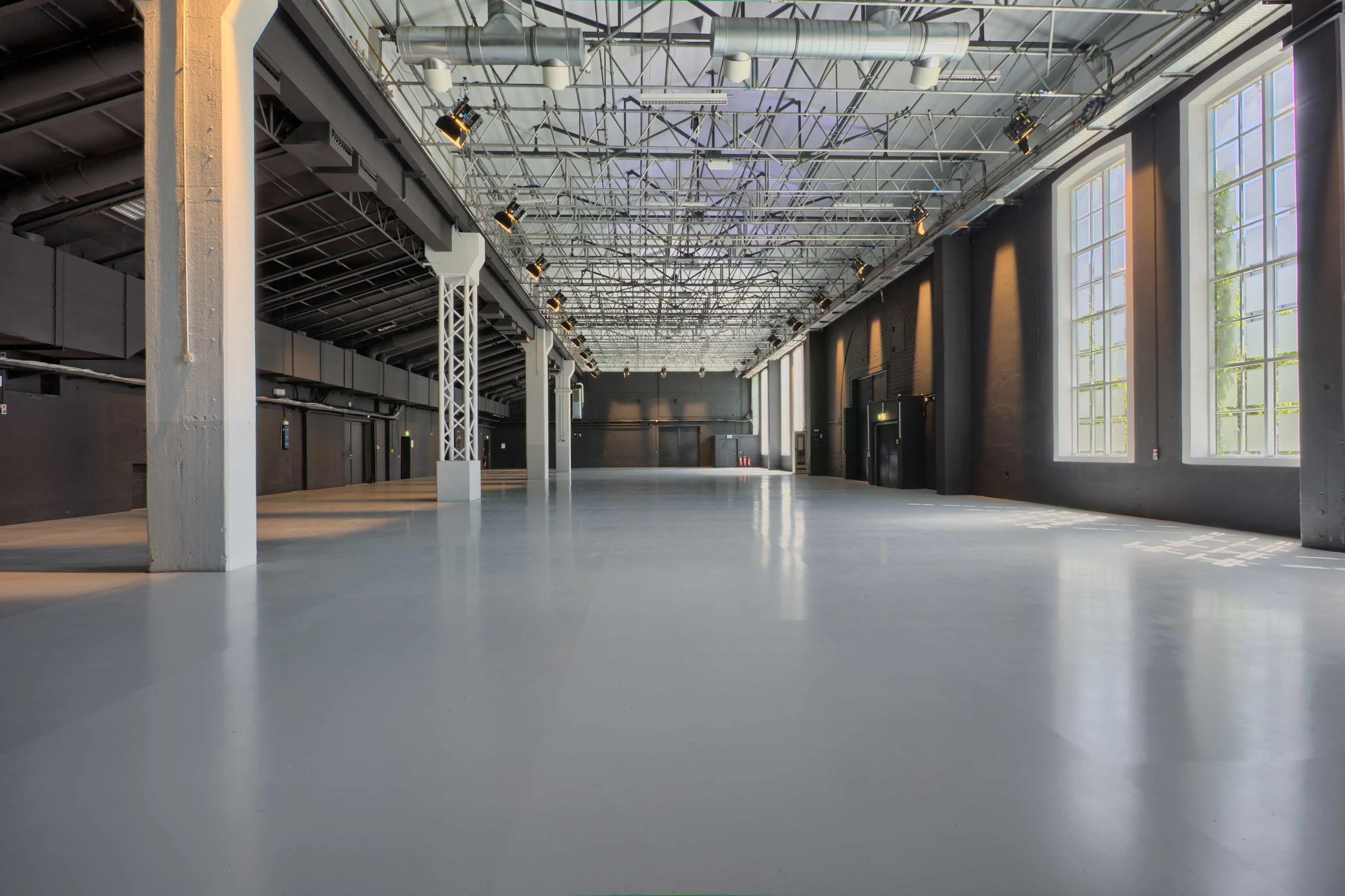 Leeres, modernes Veranstaltungsraum mit großen Fenstern, schwarzen Wänden, weißen Säulen, glatter heller Boden, Decken mit sichtbarer Stahlträger- und Lichtinstallationen.
