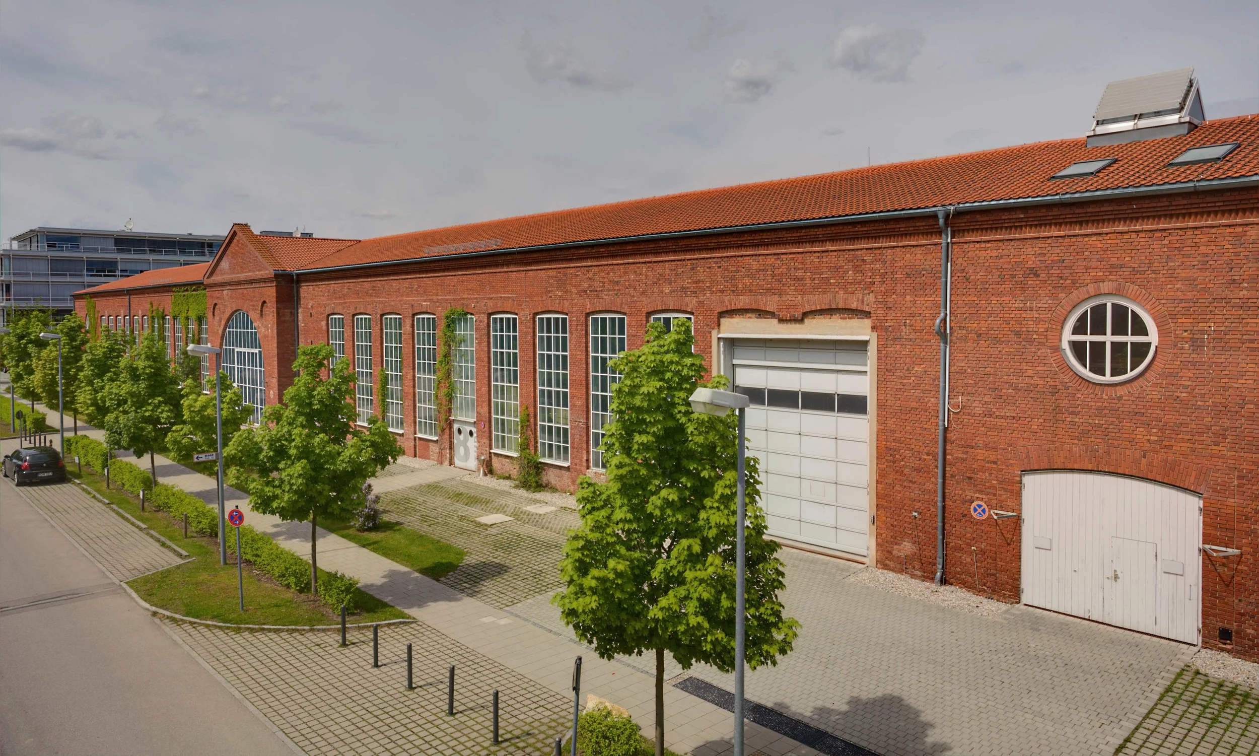 Historische rote Backstein-Fabrikhalle mit großen Fenstern, begrünten Vorplätzen und Straßen in einer urbanen Umgebung.