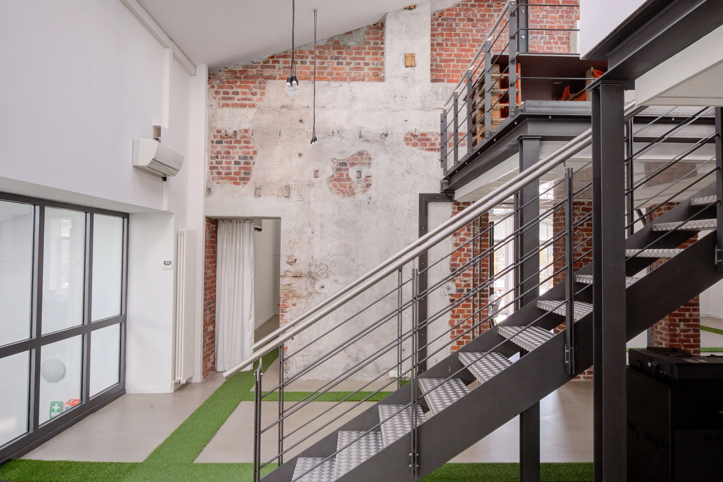 Innenansicht eines modernen, loftartigen Raumes mit offenen Steinwänden, einer schwarzen Metalltreppe und einem großen Fenster mit weißen Rahmen. Die Raumgestaltung ist industriell, mit teilweise rohem Backstein und weißen Wänden, sowie einem grünen