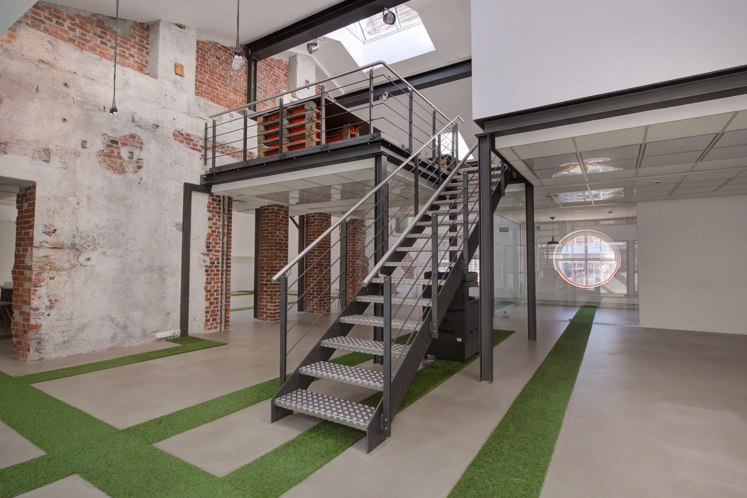 Innenaufnahme eines modernen, zweistöckigen Bürogebäudes mit offenem Grundriss, Sicht auf eine Metalltreppe, gläserne Wände, rotes Backsteinmauerwerk und künstliches Gras auf dem Boden.