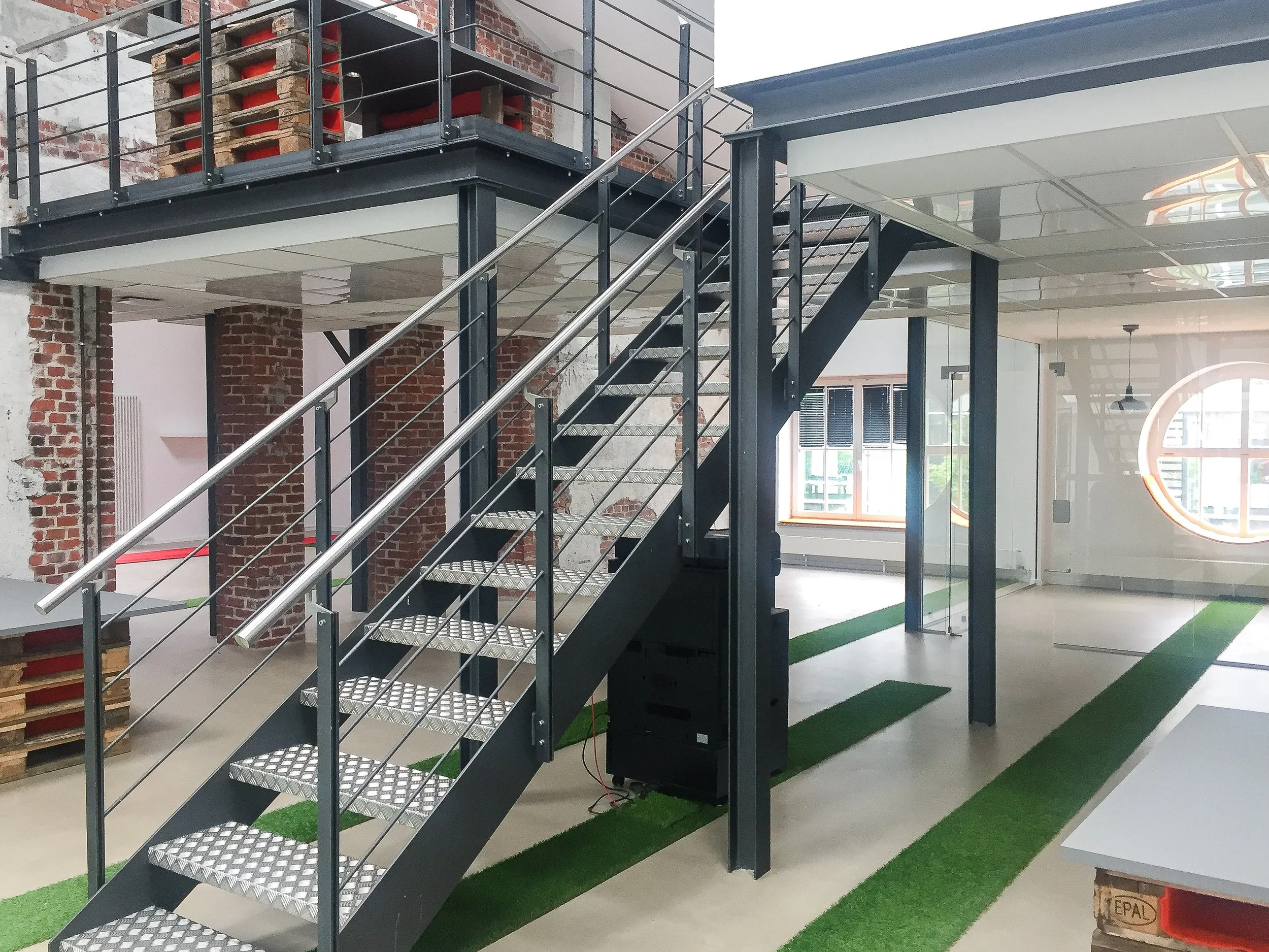 Innenansicht eines modernen, offenen Bürogebäudes mit schwarzer Metalltreppe und Brüstung, massiven Ziegelwänden im Hintergrund und großen Fenstern, die Tageslicht hereinlassen.