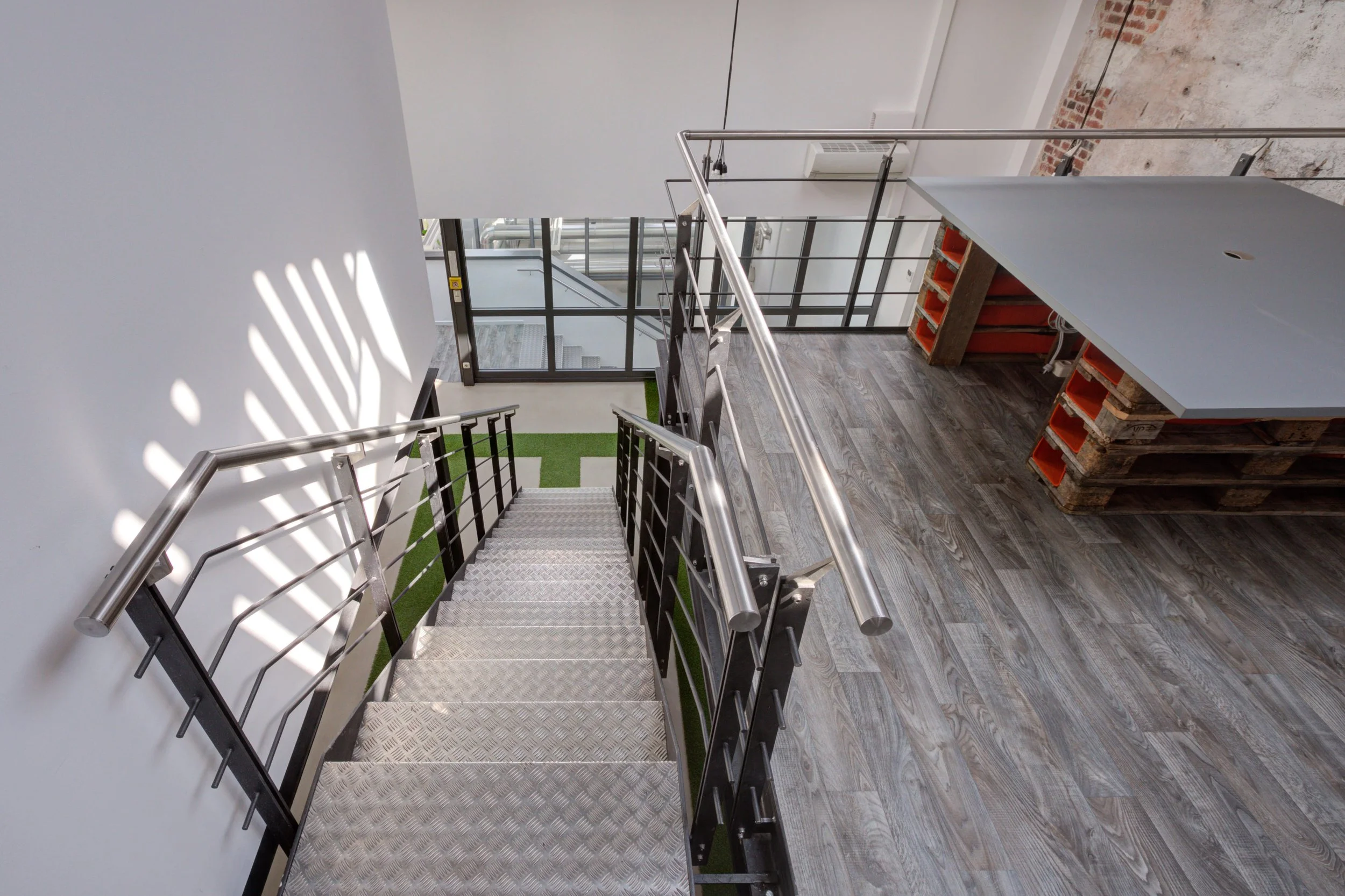 Innenaufnahme einer Treppe mit Metallhandlauf, Blick nach unten auf eine Etage mit Holzboden und einem Tisch aus Paletten, im Hintergrund große Fenster und eine Tür.