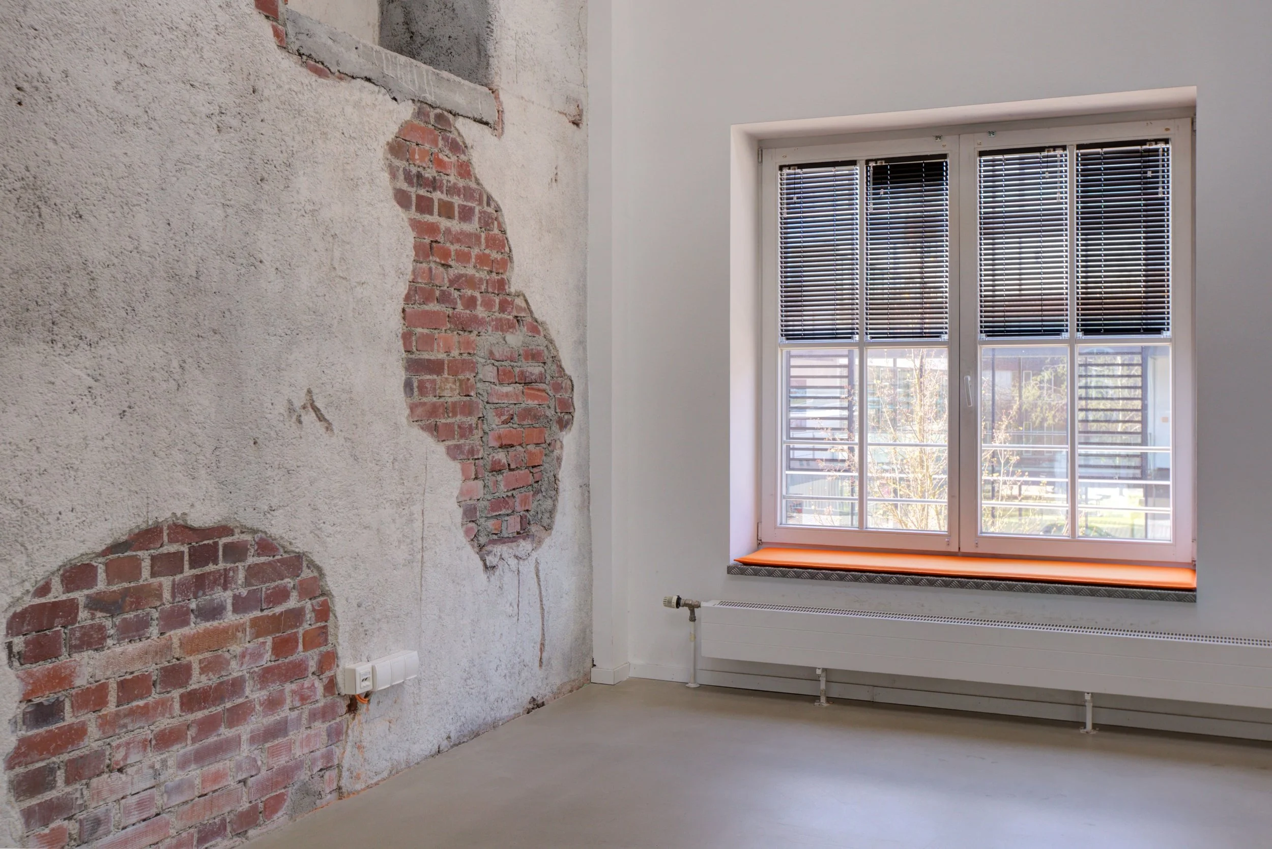 Innenraum eines Zimmers mit einer Fensterscheibe, die Rollläden hat; die Wand hat absehbare Stellen mit sichtbaren Ziegelsteinen und Putz