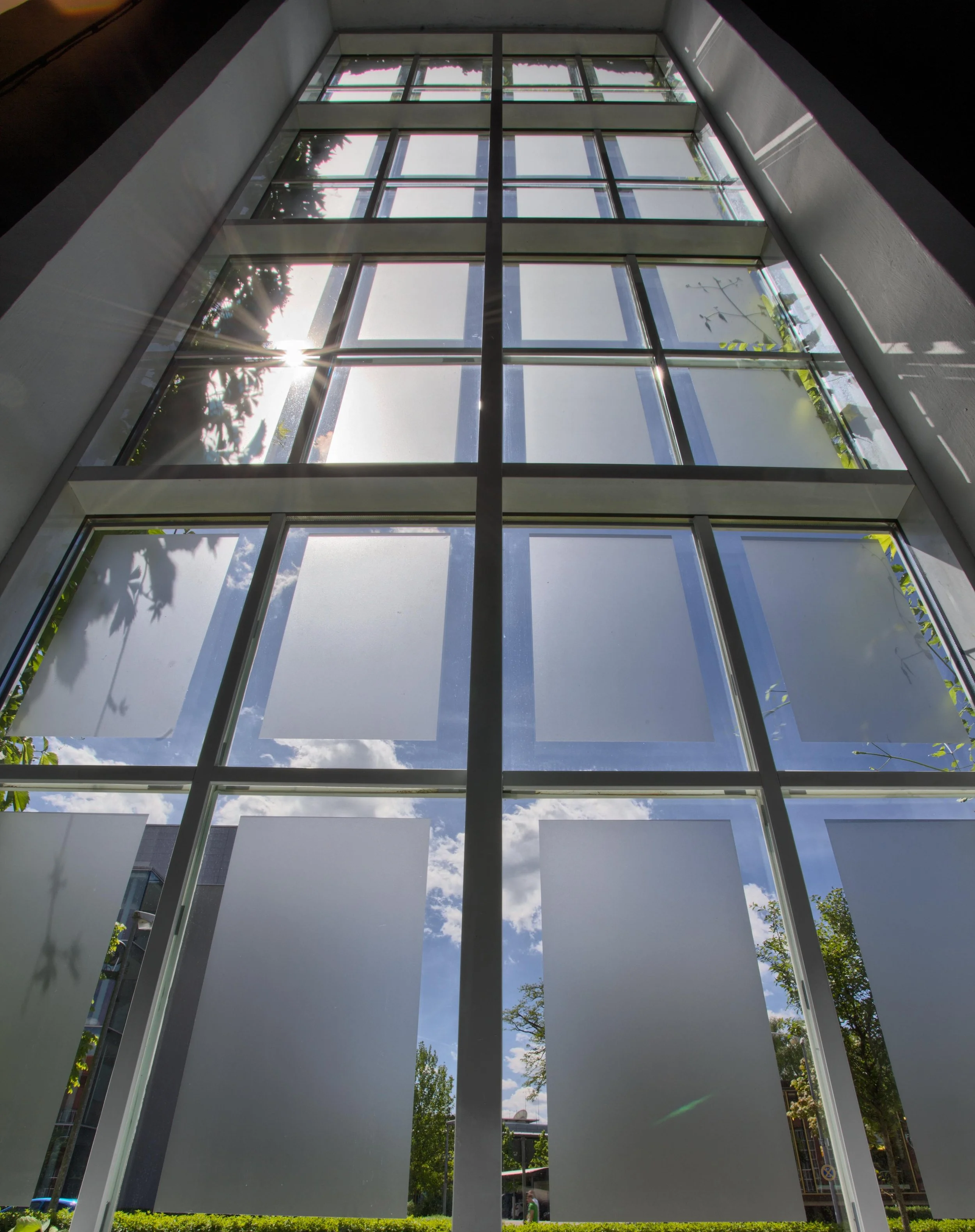 Blick nach oben auf ein großes Glasfenster in einem modernen Gebäude, durch das der Himmel mit Wolken und Sonnenschein sichtbar ist.