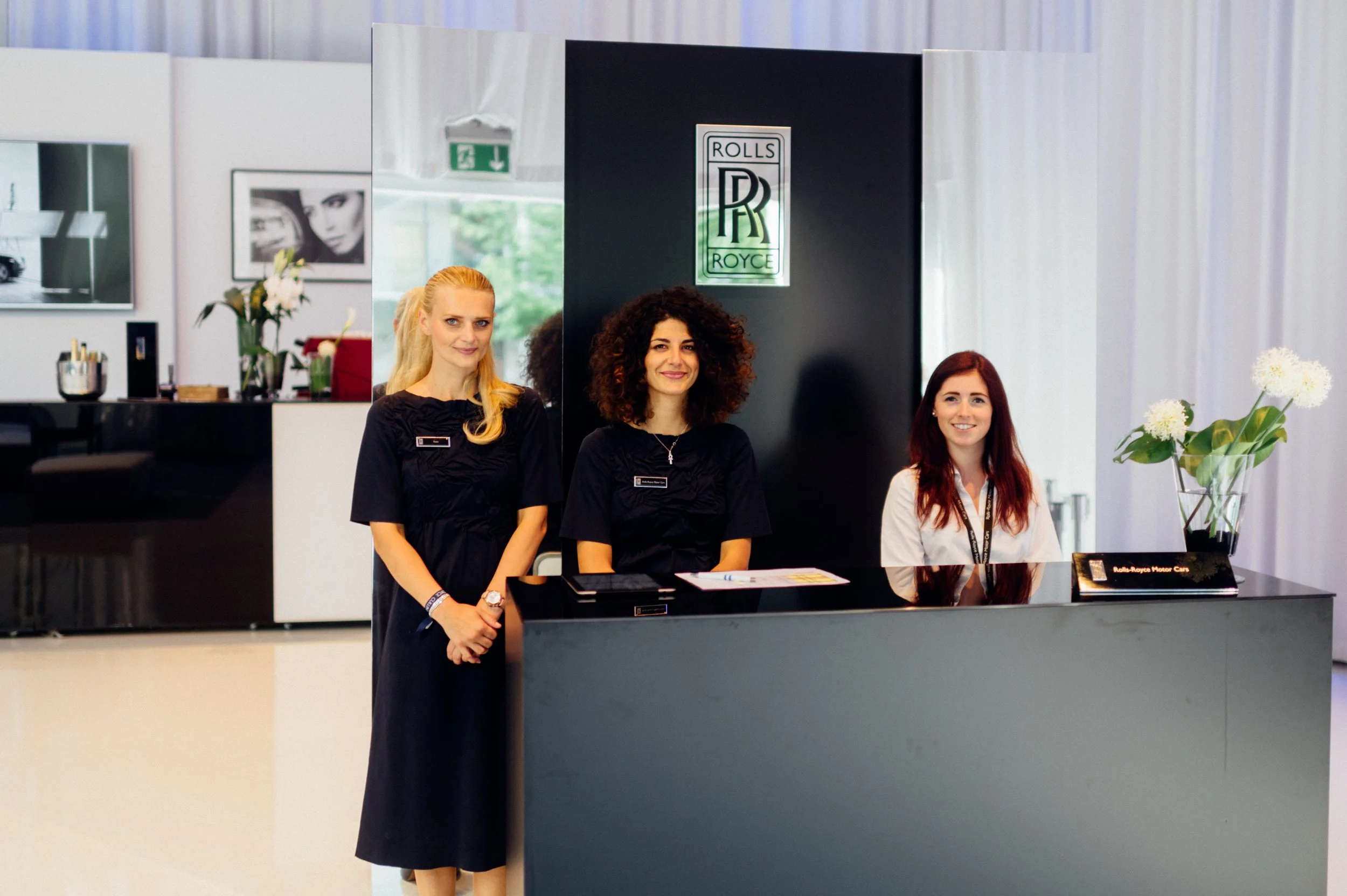 Drei Damen stehen hinter einem Empfangstresen im Autohaus, im Hintergrund sind Blumen und Bilder zu sehen.