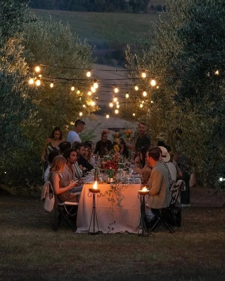 A group of people are gathered around a long table outdoors during the evening, with string lights overhead creating a warm ambiance. The table is decorated with flowers, and there are candles and drinks on the table. The setting appears to be a dinner party or celebration in a natural, scenic environment surrounded by trees and greenery.