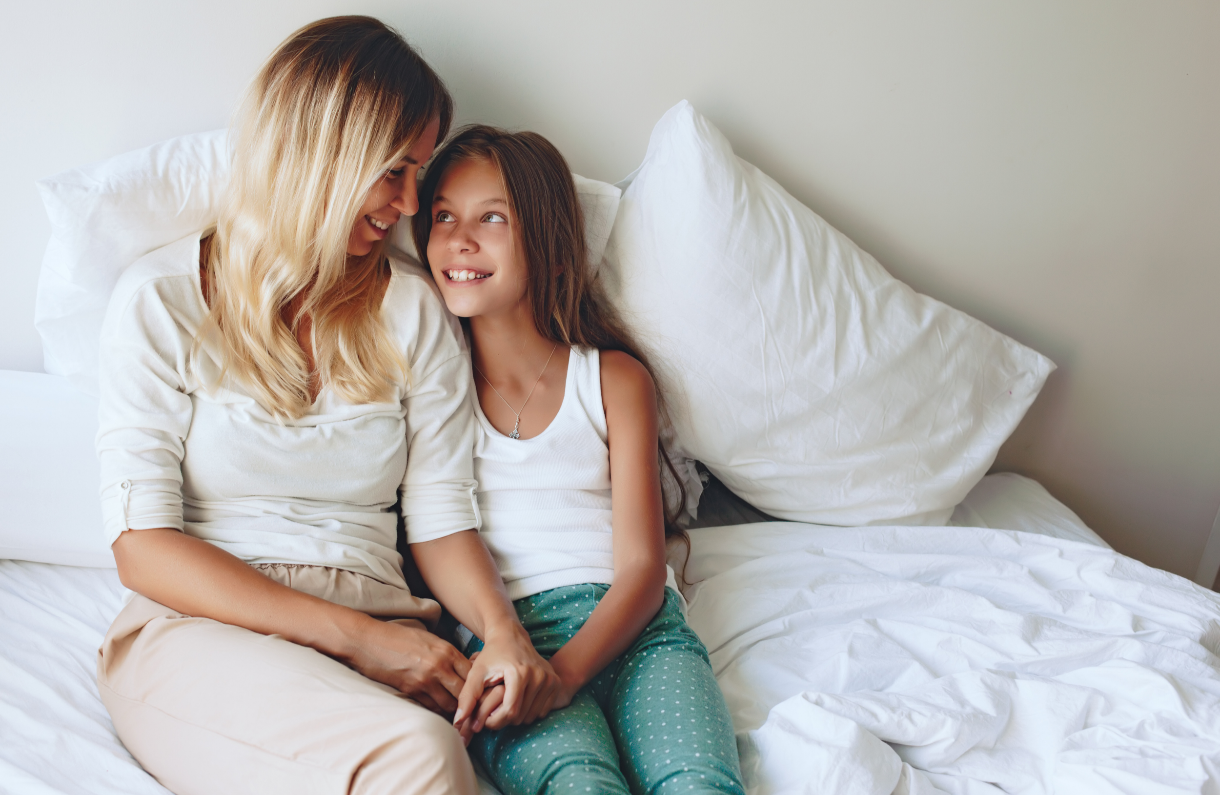A woman and a young girl sitting on a bed, smiling and looking at each other.