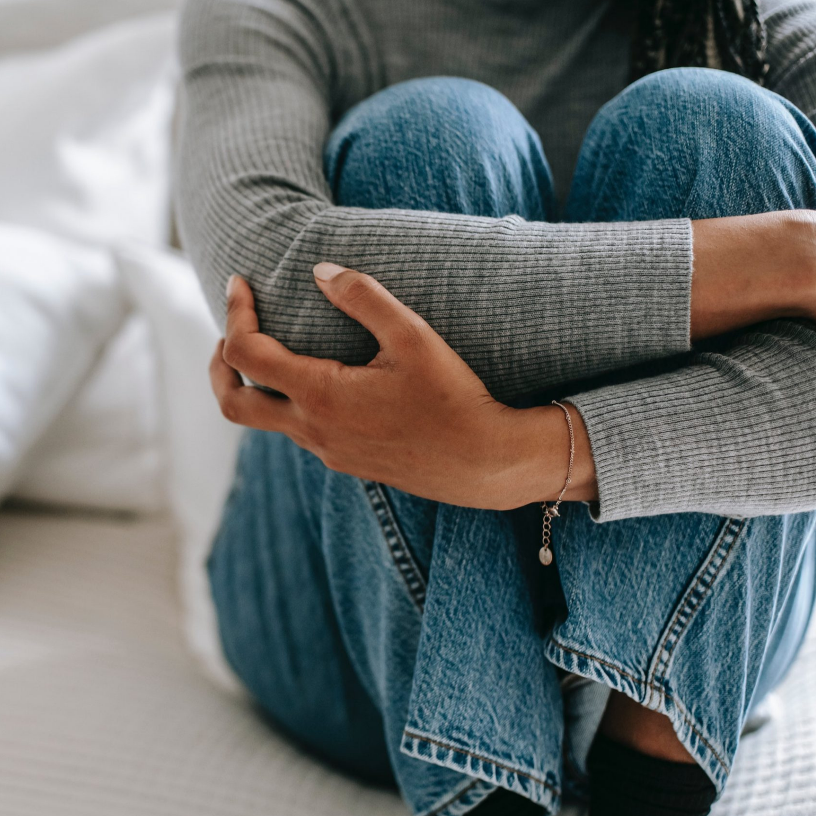 Person sitting on a bed, hugging their knees, wearing a gray ribbed sweater and blue jeans, with a silver bracelet on their wrist.
