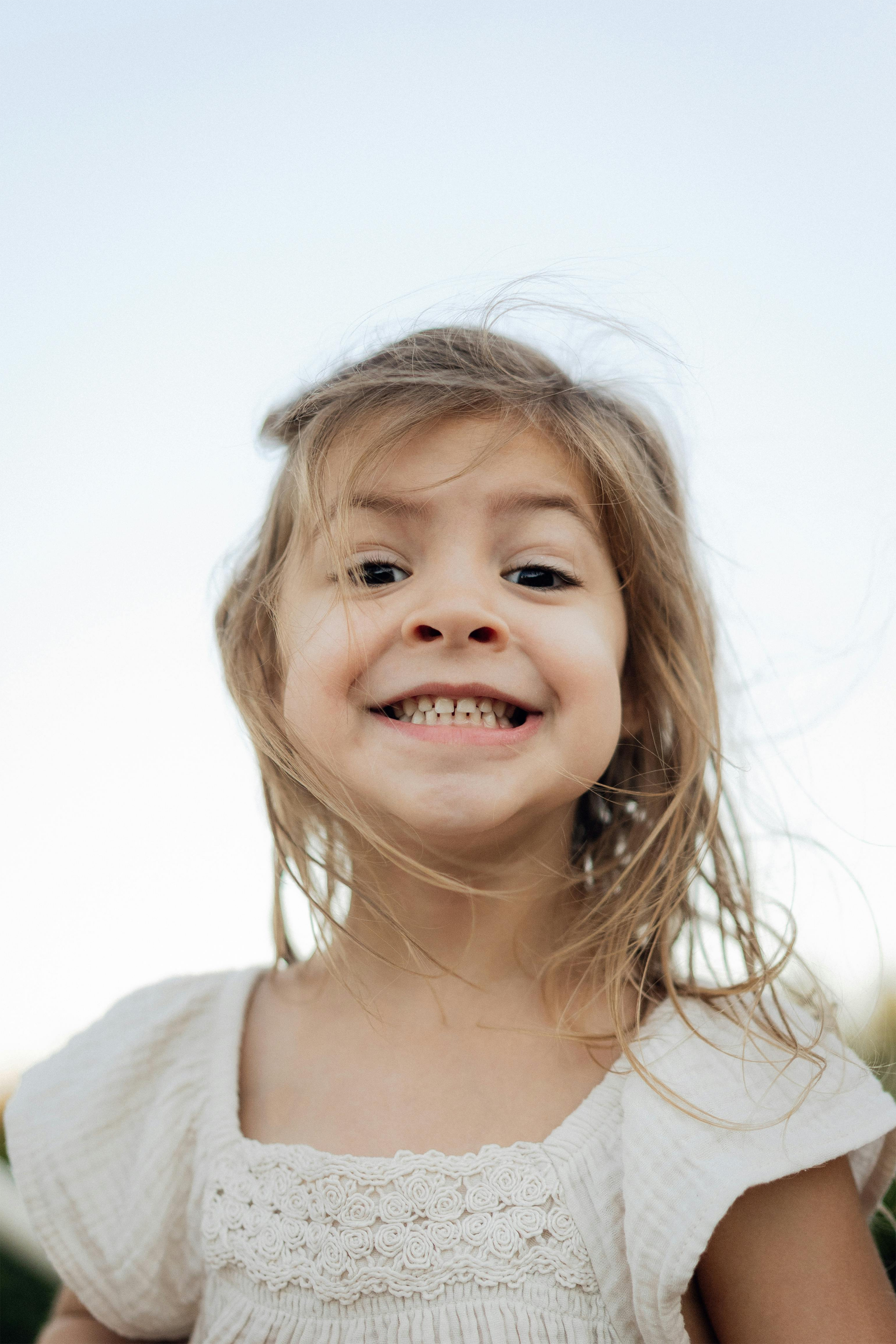 A young girl with light brown hair smiling at the camera, wearing a white dress with embroidered details, outdoors against a light sky background.