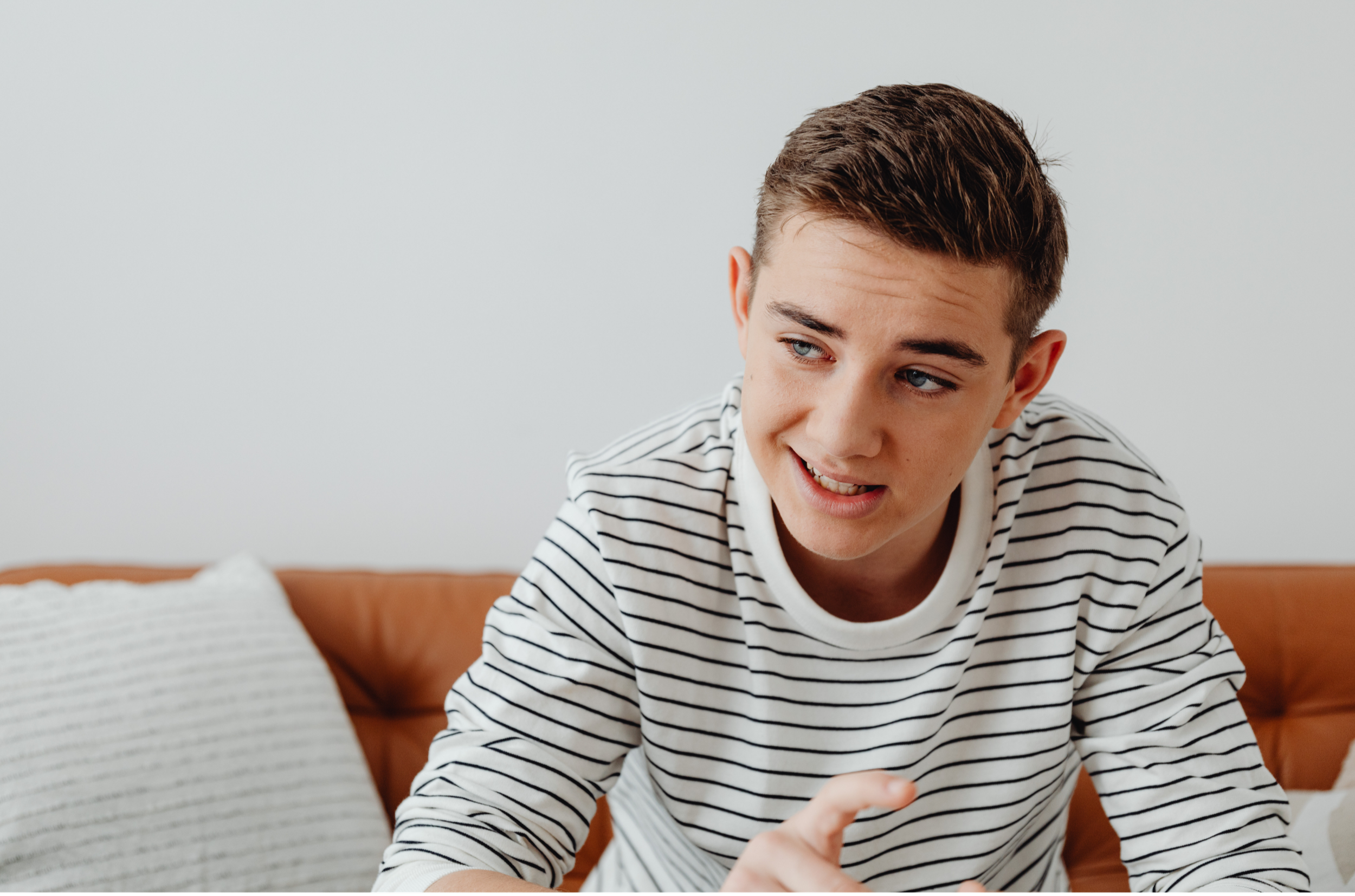 A young man with short brown hair and blue eyes wearing a white and black striped shirt, sitting on a brown leather couch in a room with a white wall, engaged in a conversation.