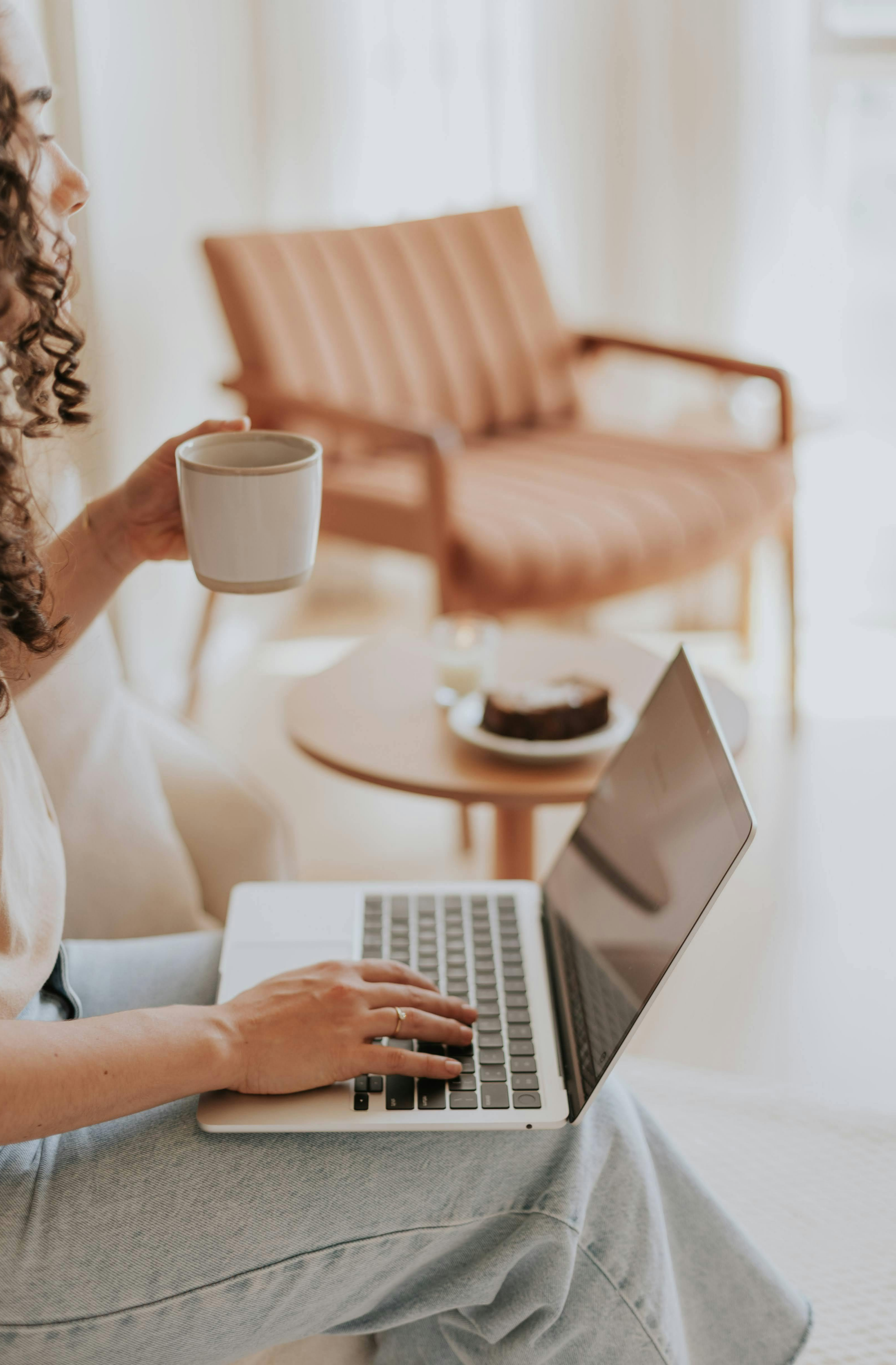 A woman sitting on a beige sofa, using a laptop with a cup in her hand, in a bright living room with a side table holding a plate and a dessert.