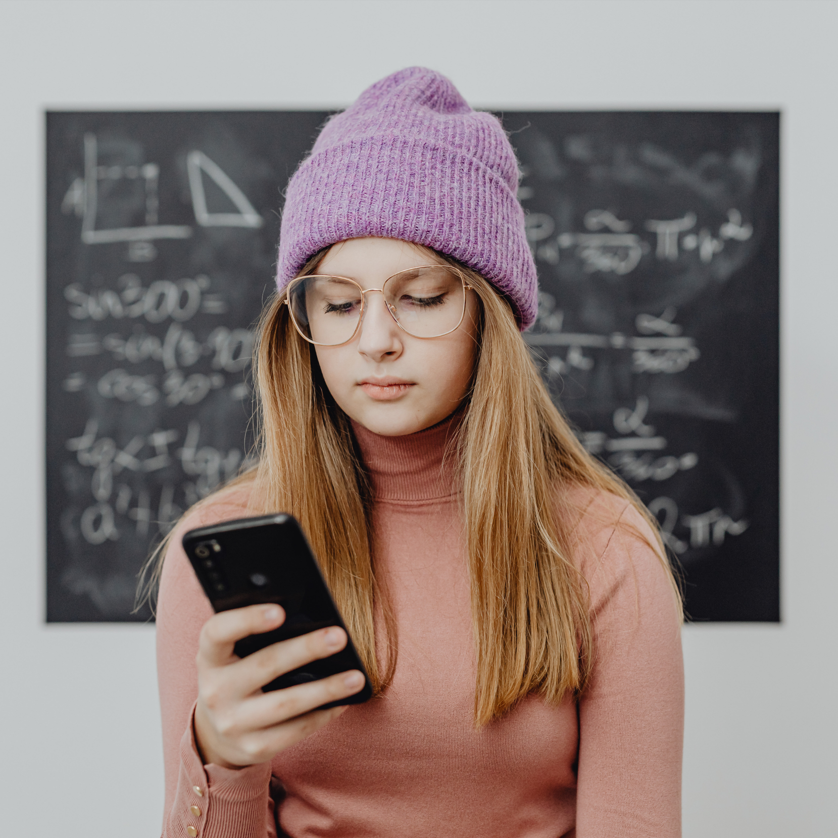 A young woman with long blonde hair, wearing a pink turtleneck sweater, a purple knit hat, and glasses, looking at her smartphone in a classroom with a blackboard filled with mathematical equations in the background.