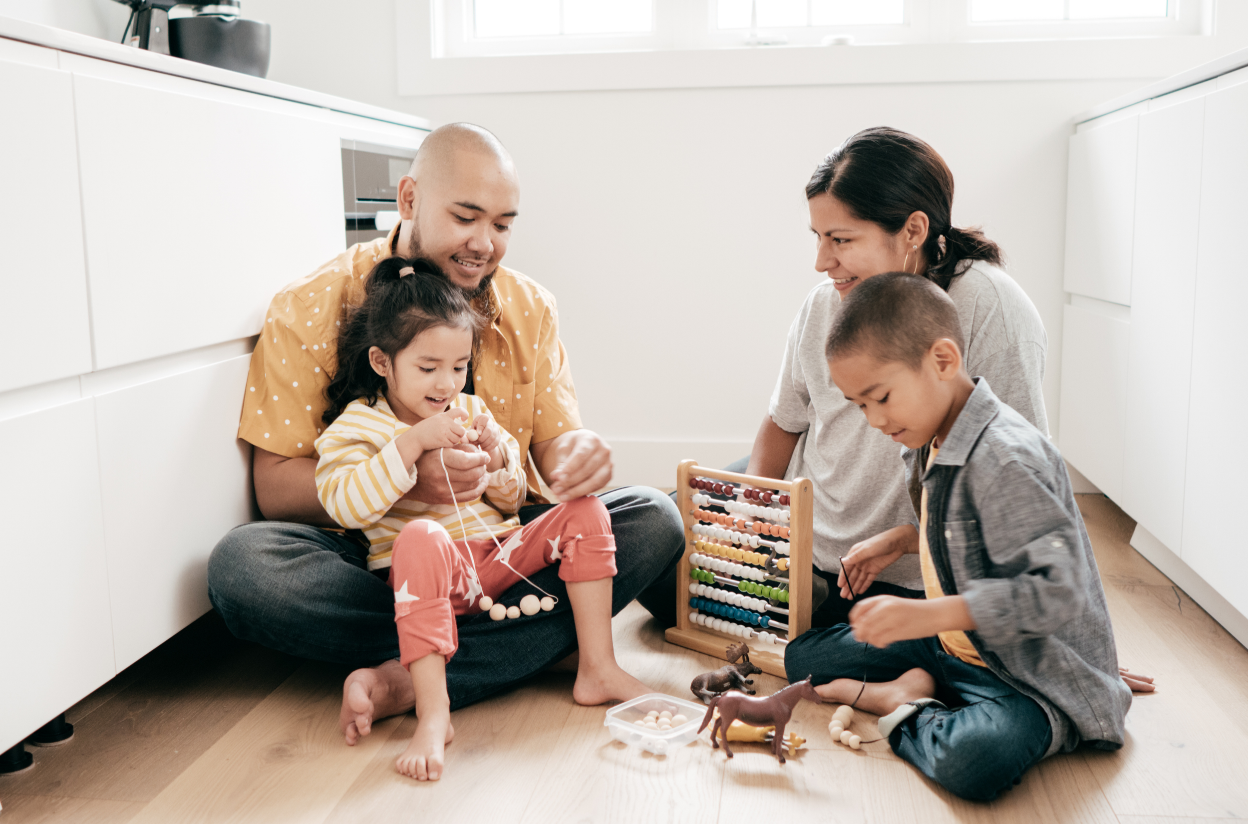 Family of four, two adults and two children, playing with toys on the floor of a bright, modern kitchen.