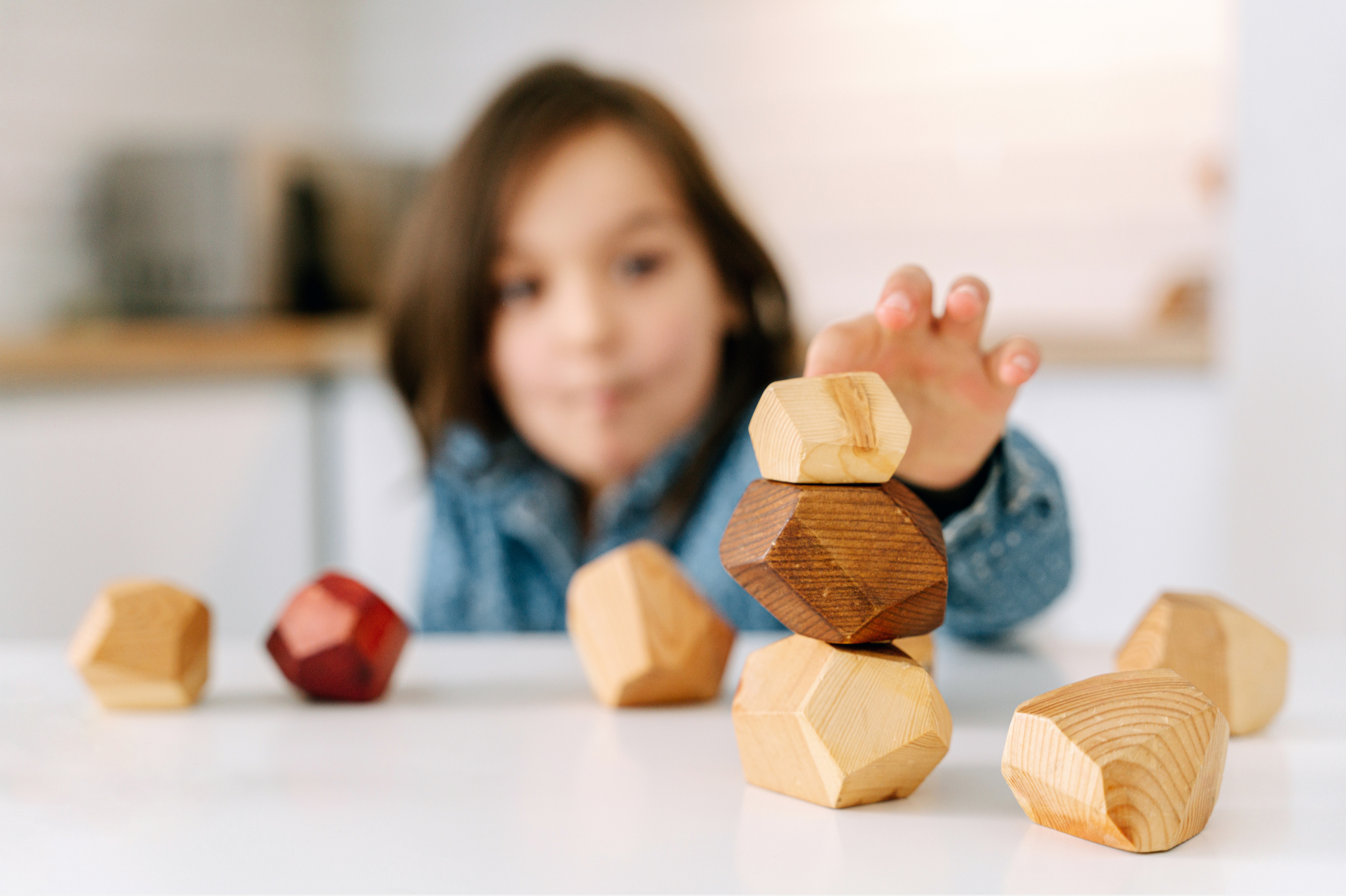 Child stacking wooden geometric blocks on a table with several more blocks scattered around.
