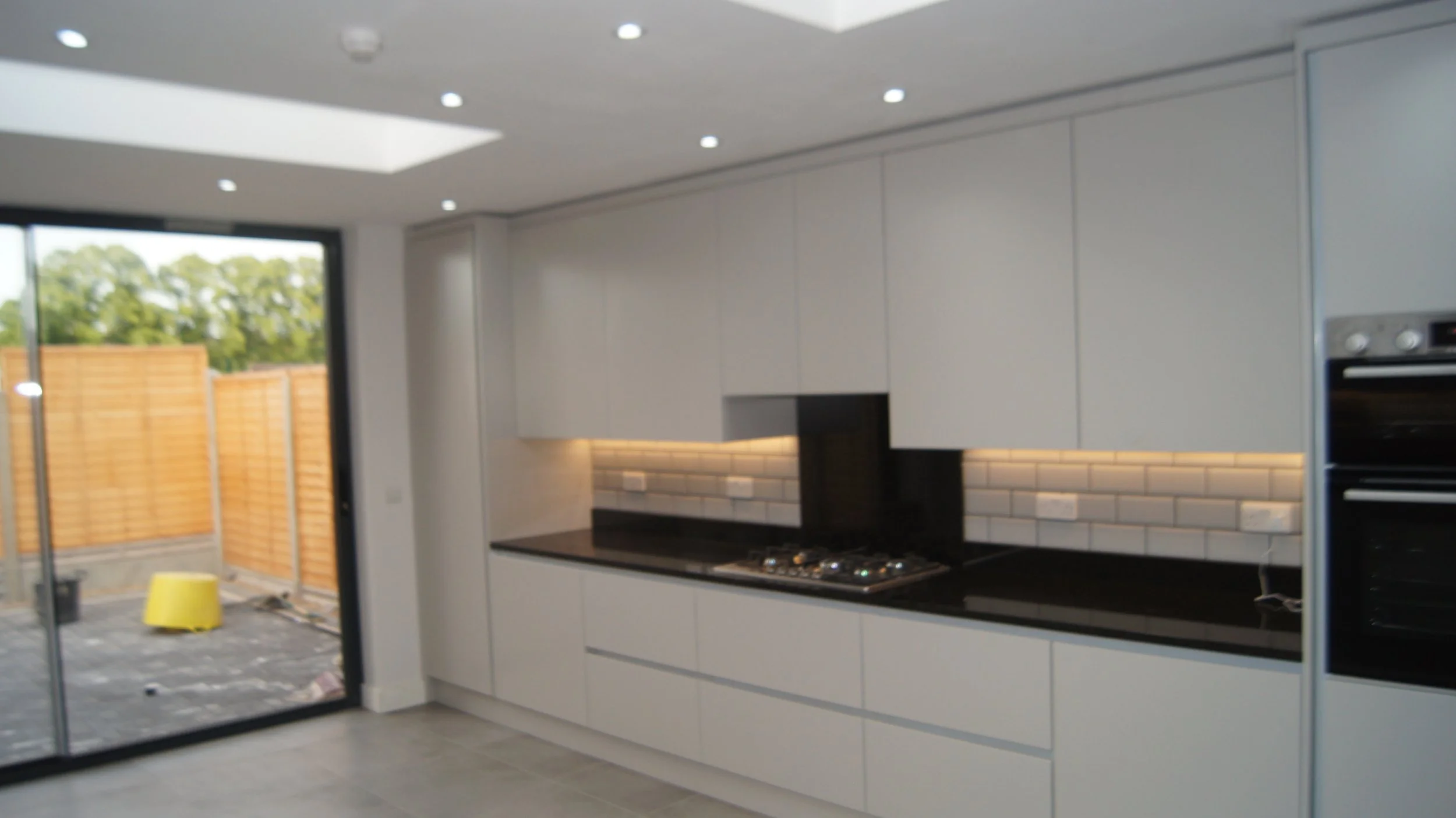 Modern kitchen with white cabinets, black countertop, white subway tile backsplash, gas stove, and built-in oven. Large sliding glass door opens to outdoor patio with wooden fencing and green trees.