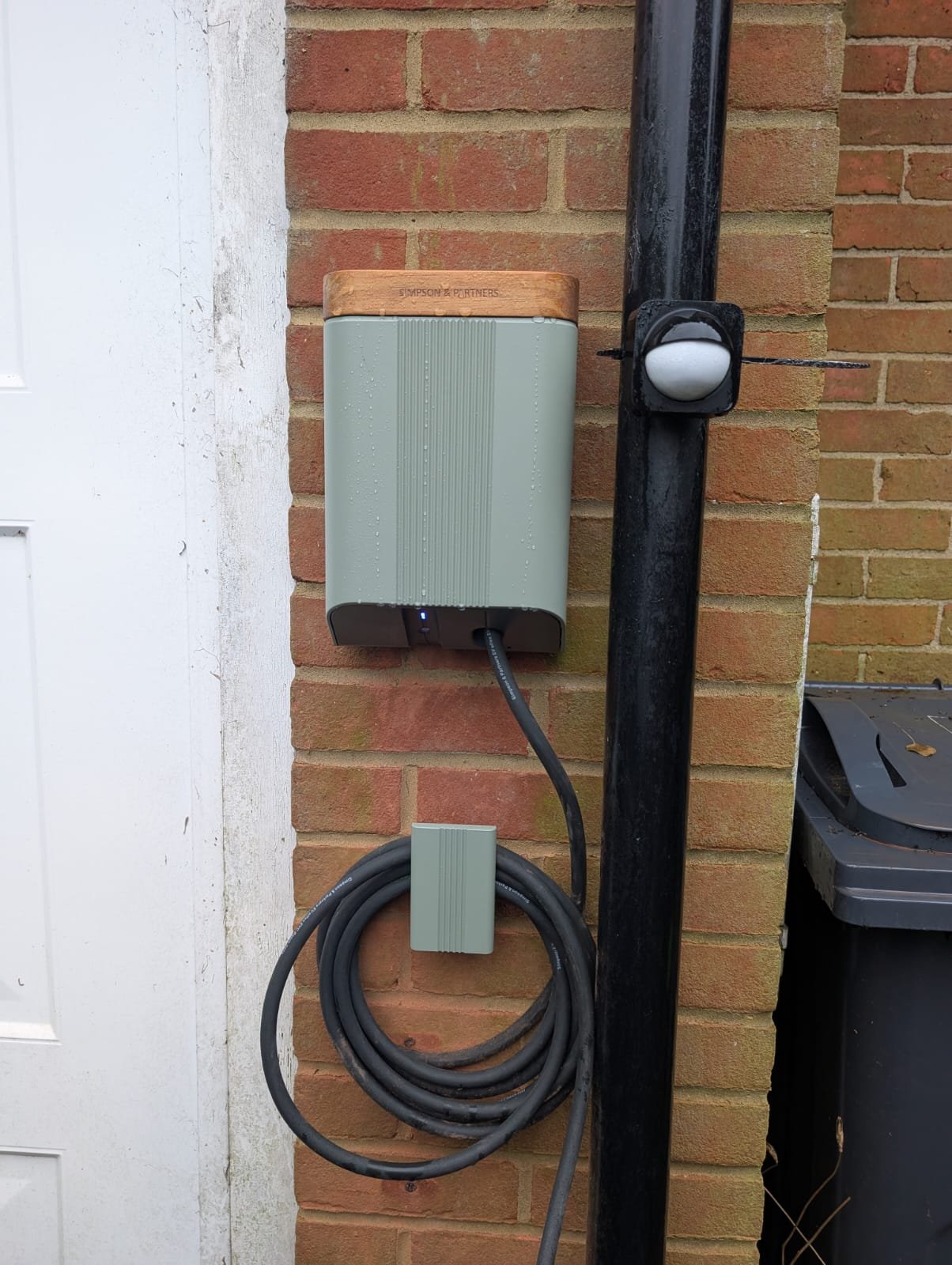 Outdoor electrical charging station mounted on a brick wall, with a coiled cord hanging below it and a black post with a switch or outlet to the right.