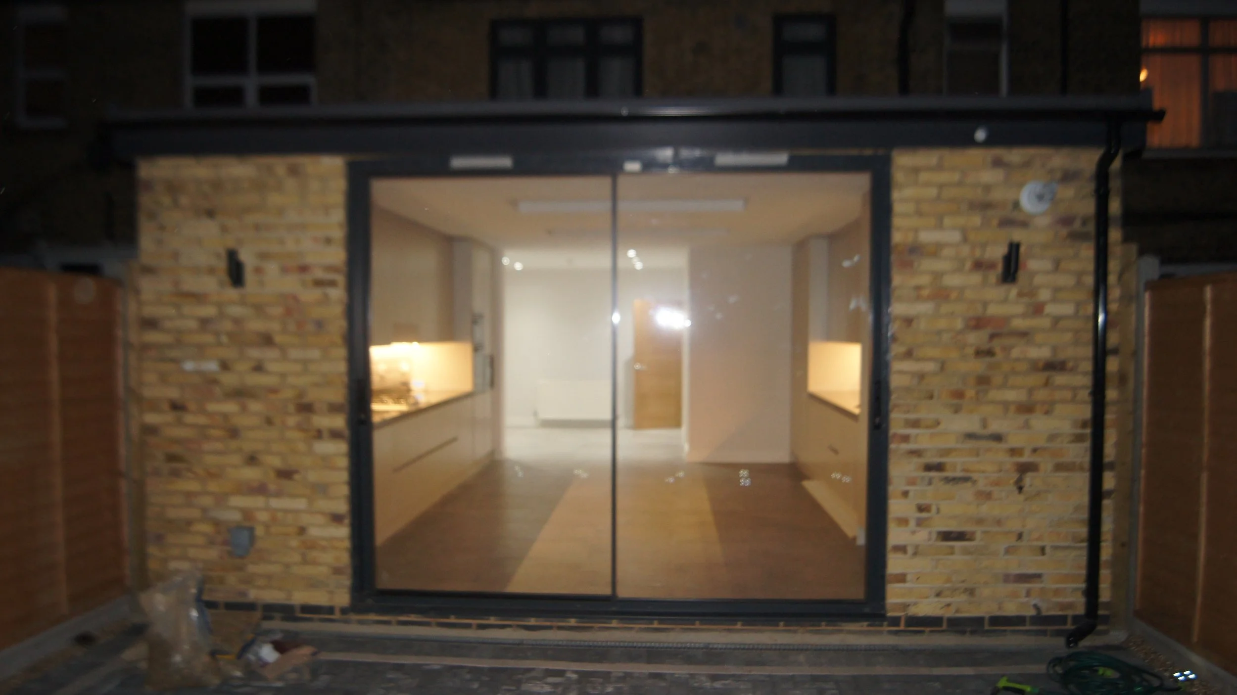 View of a glass door opening into an interior space with white walls and a wooden floor, enclosed by a brick exterior structure.