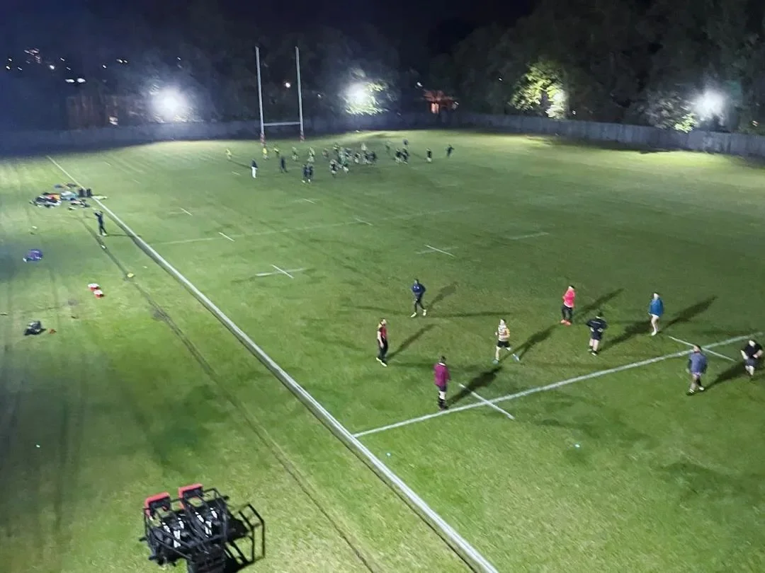 A nighttime scene at a sports field with players practicing or warming up while a group sits or stands in the background. Bright floodlights illuminate the field.